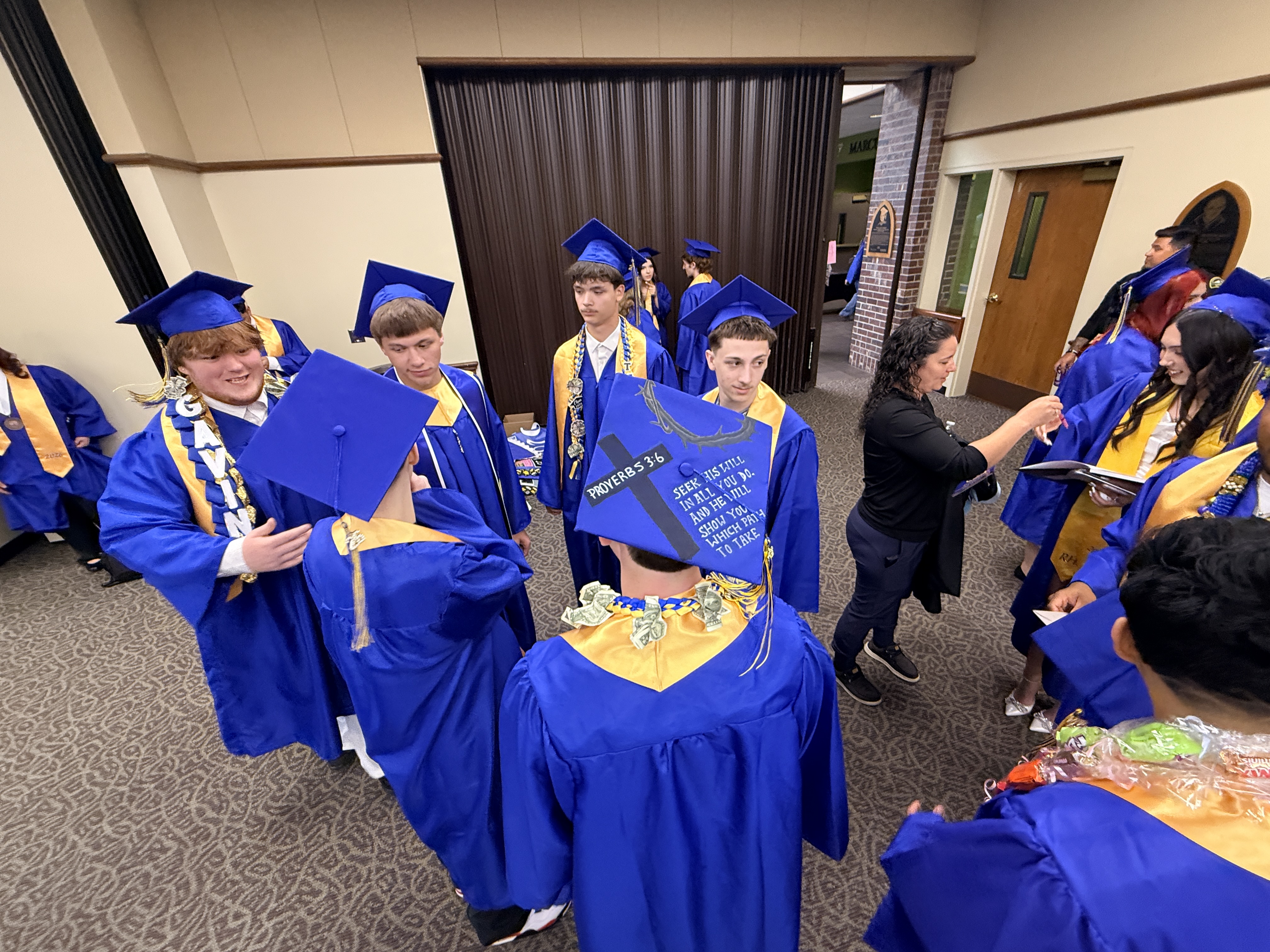 Graduates in blue and gold caps and gowns gather in a room with a brown door.