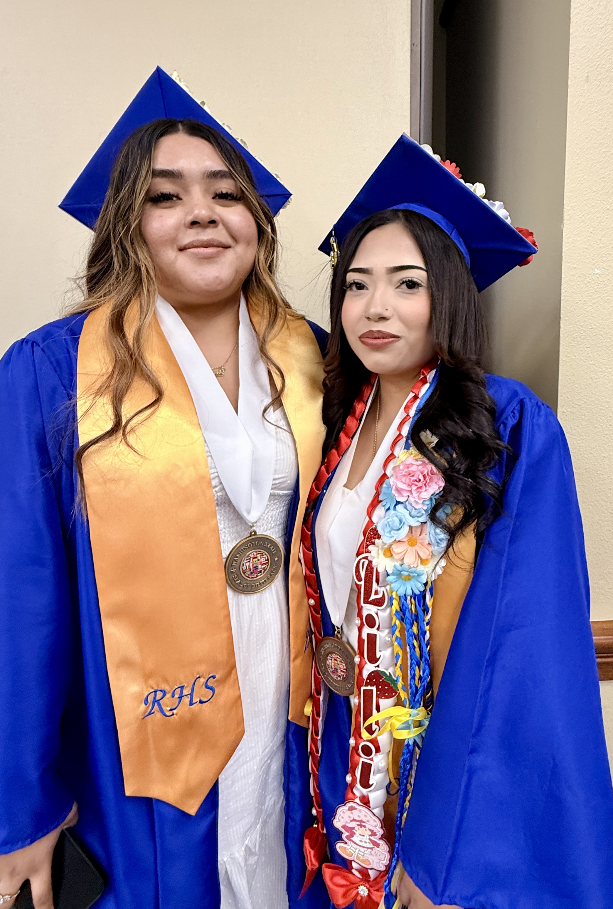 Two women in blue graduation gowns, one with a sash and medal, stand next to each other, smiling.