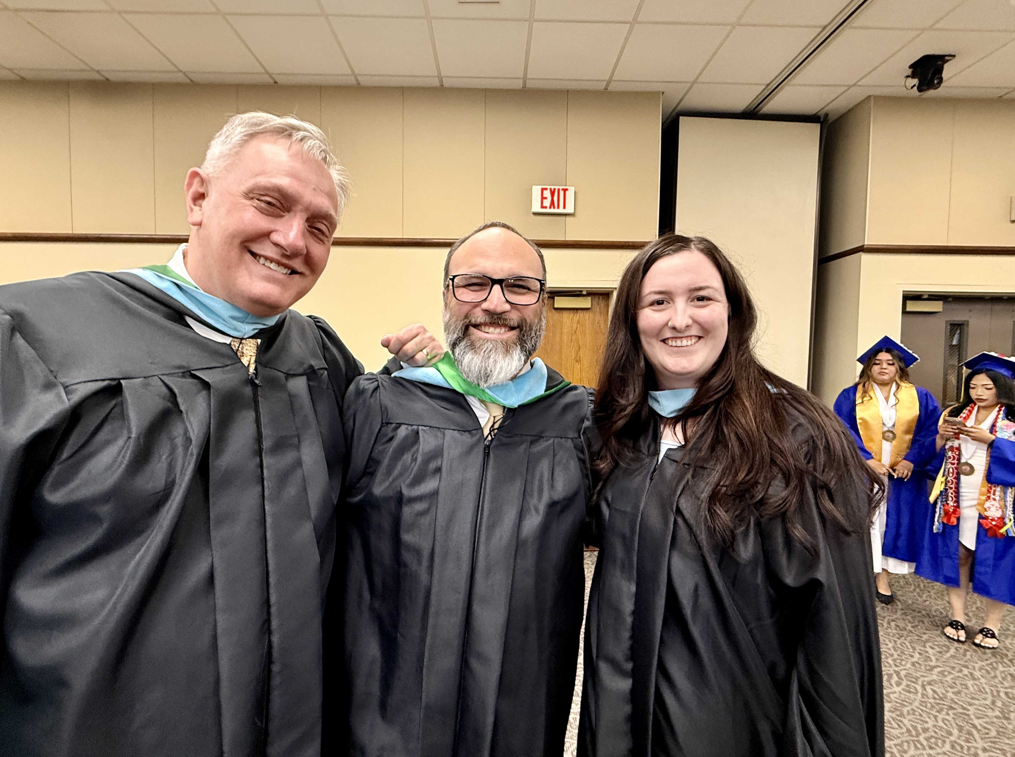 Three individuals in academic gowns and caps pose for a photo. Behind them, additional people in graduation attire are visible.
