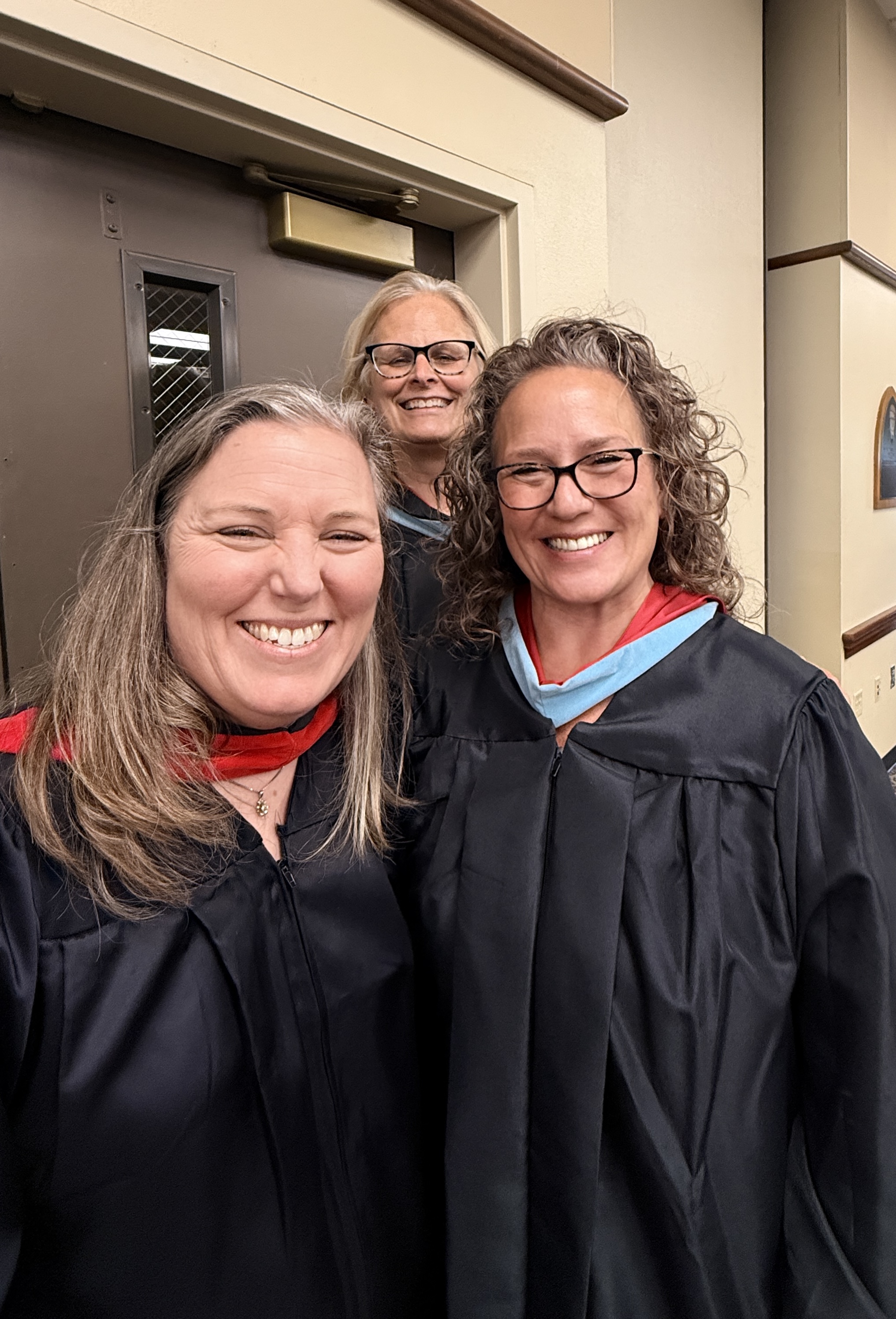 Three women in black graduation gowns smile and stand together. A door and wall with a window are in the background.