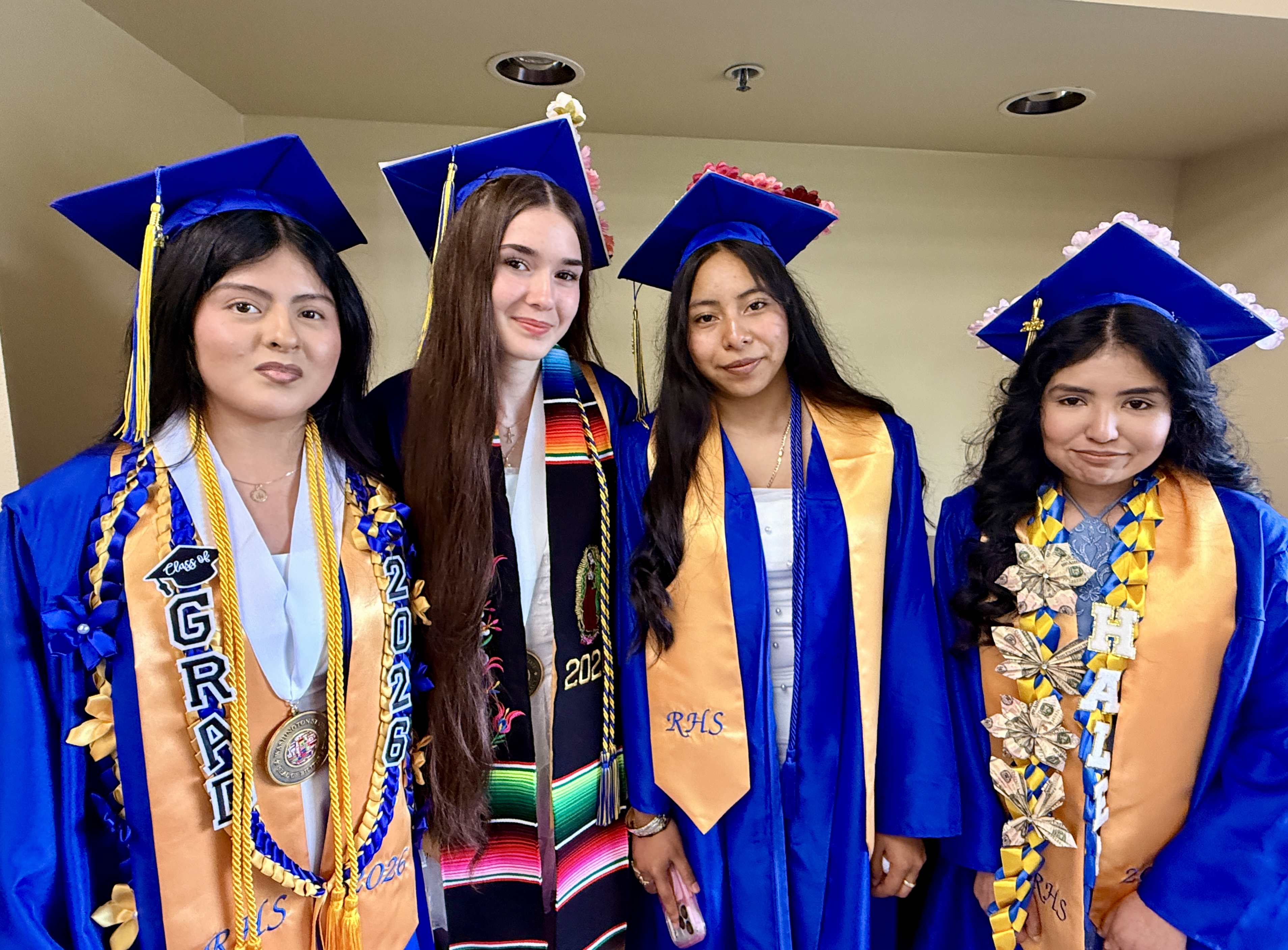 Five women in graduation gowns, one with a sash, stand together, smiling for a photo.