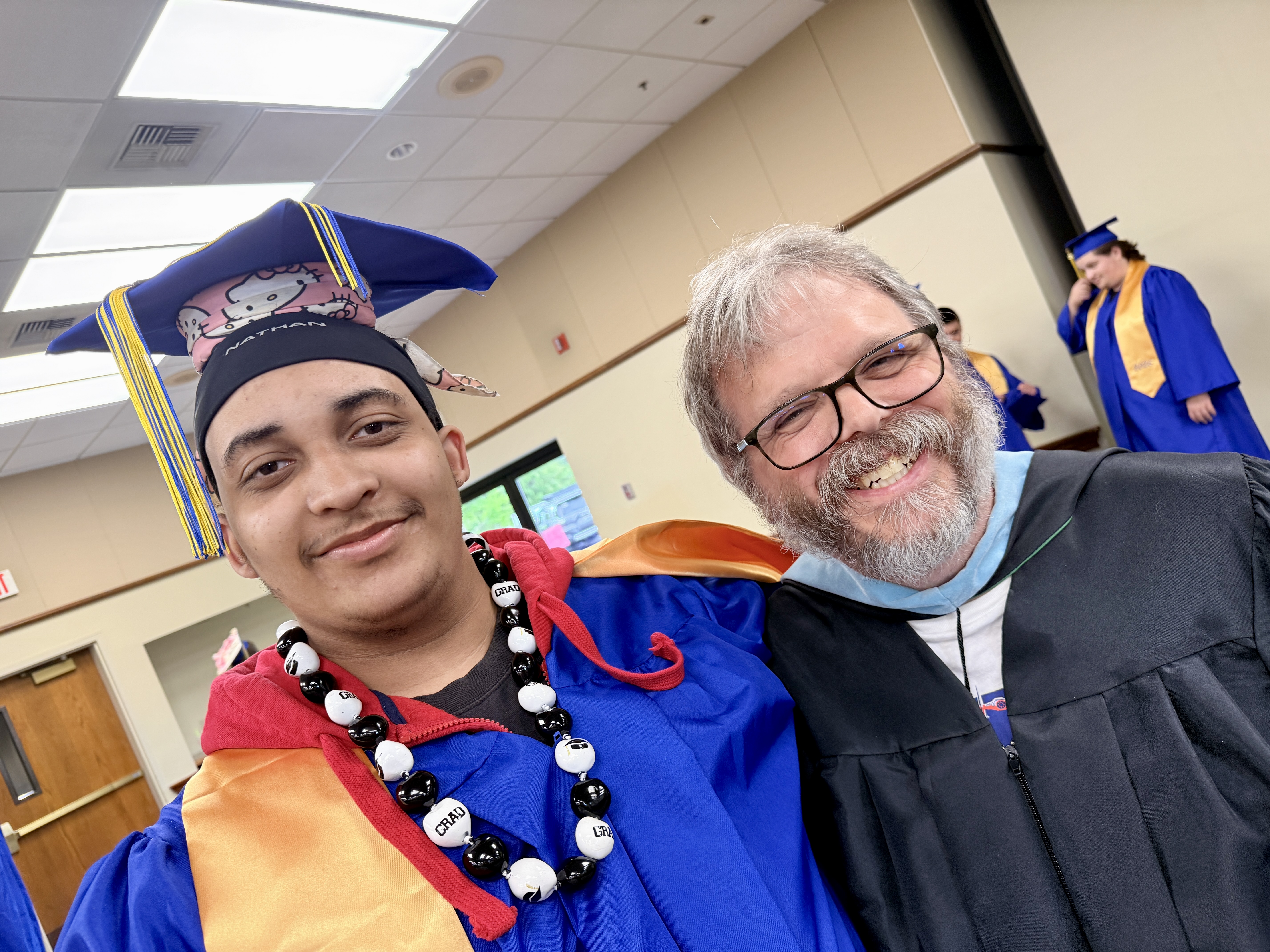 Two men, one wearing a cap and gown, the other in glasses and a graduation robe, smile for the camera.
