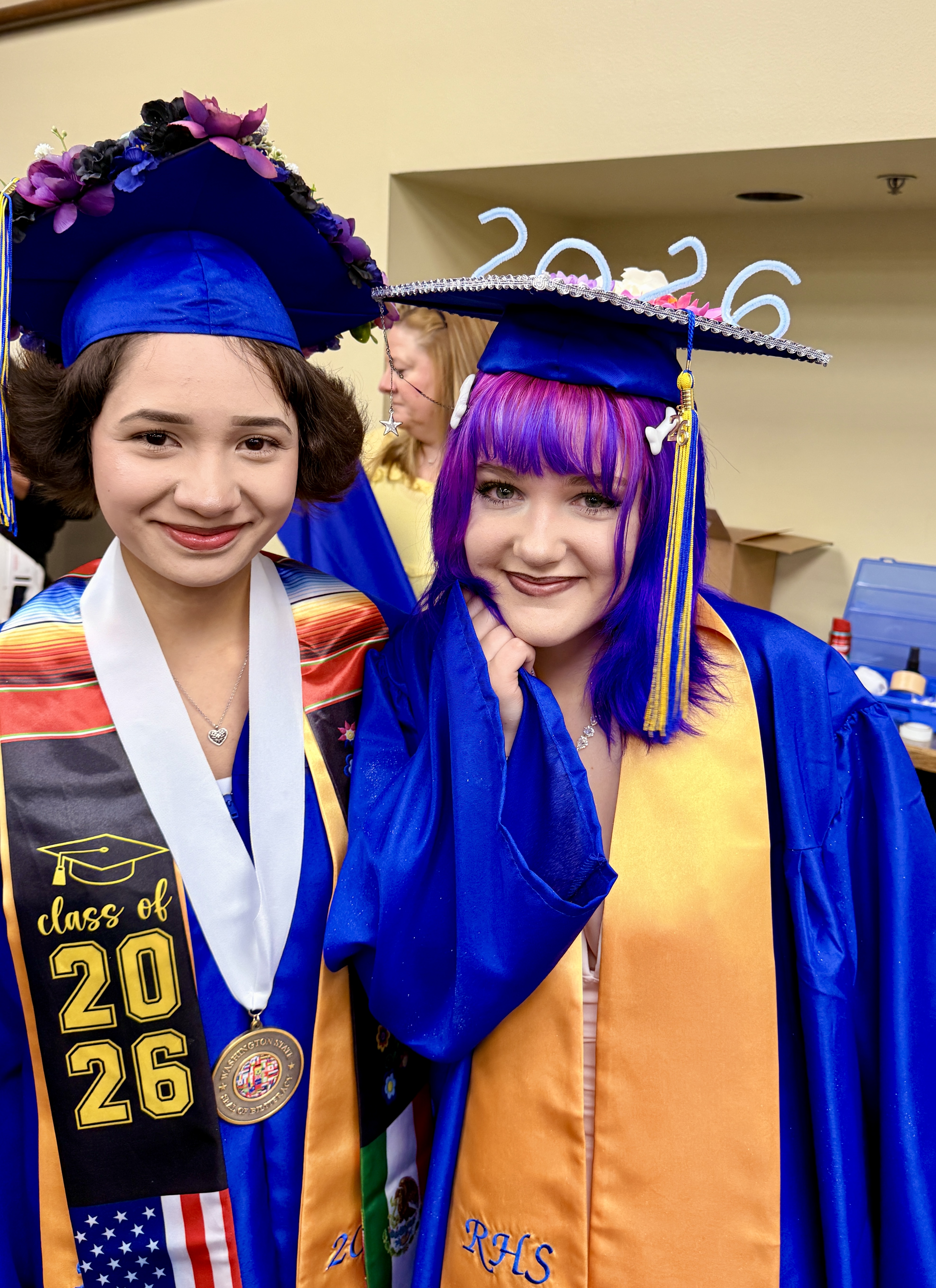Two women in graduation gowns and hats. One wears a sash with "Class of 2026" and an American flag, the other has purple hair.