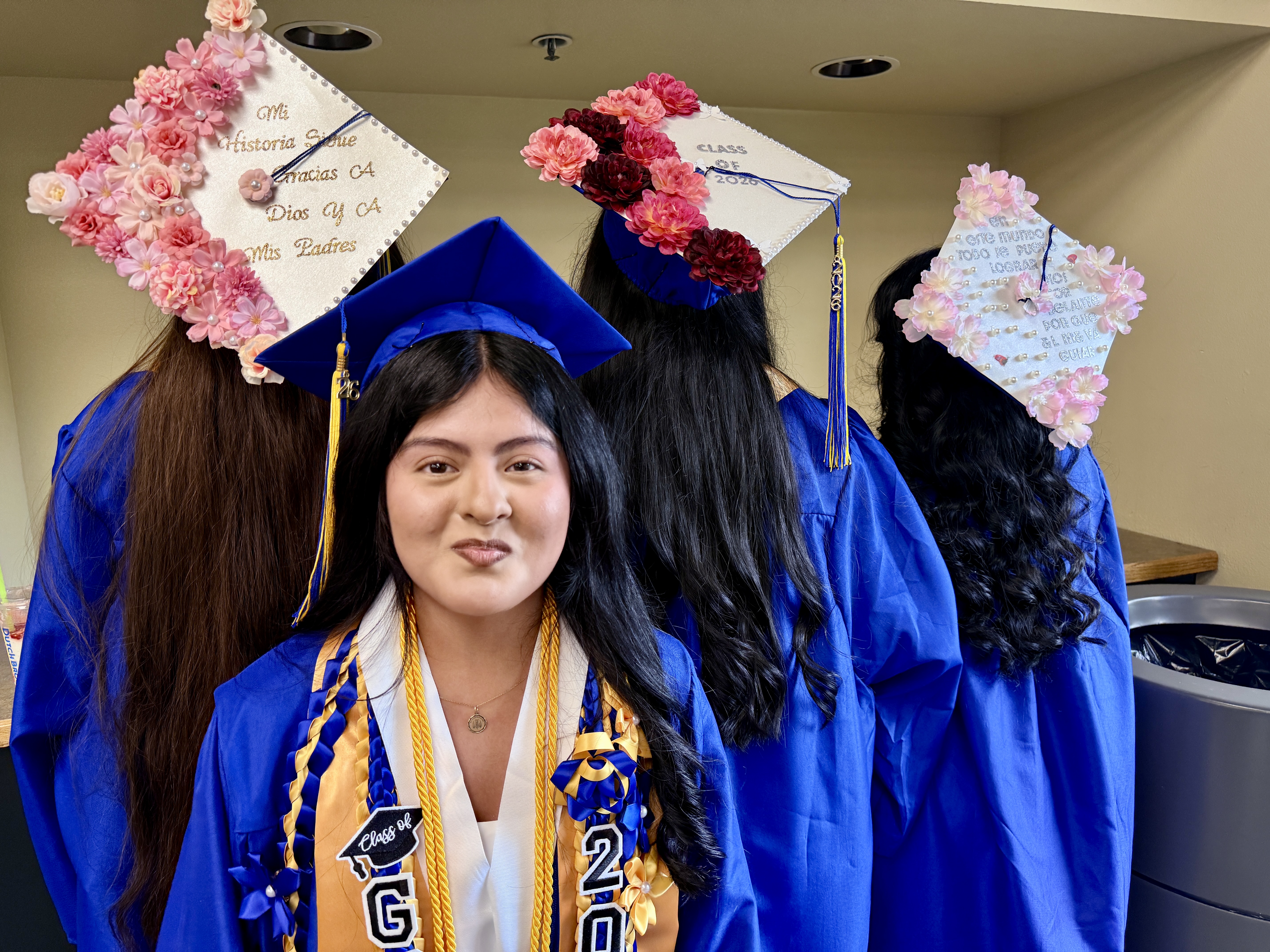 Graduates in blue gowns and caps, adorned with flowers, smile for a picture. One woman has a cap that reads "2020".