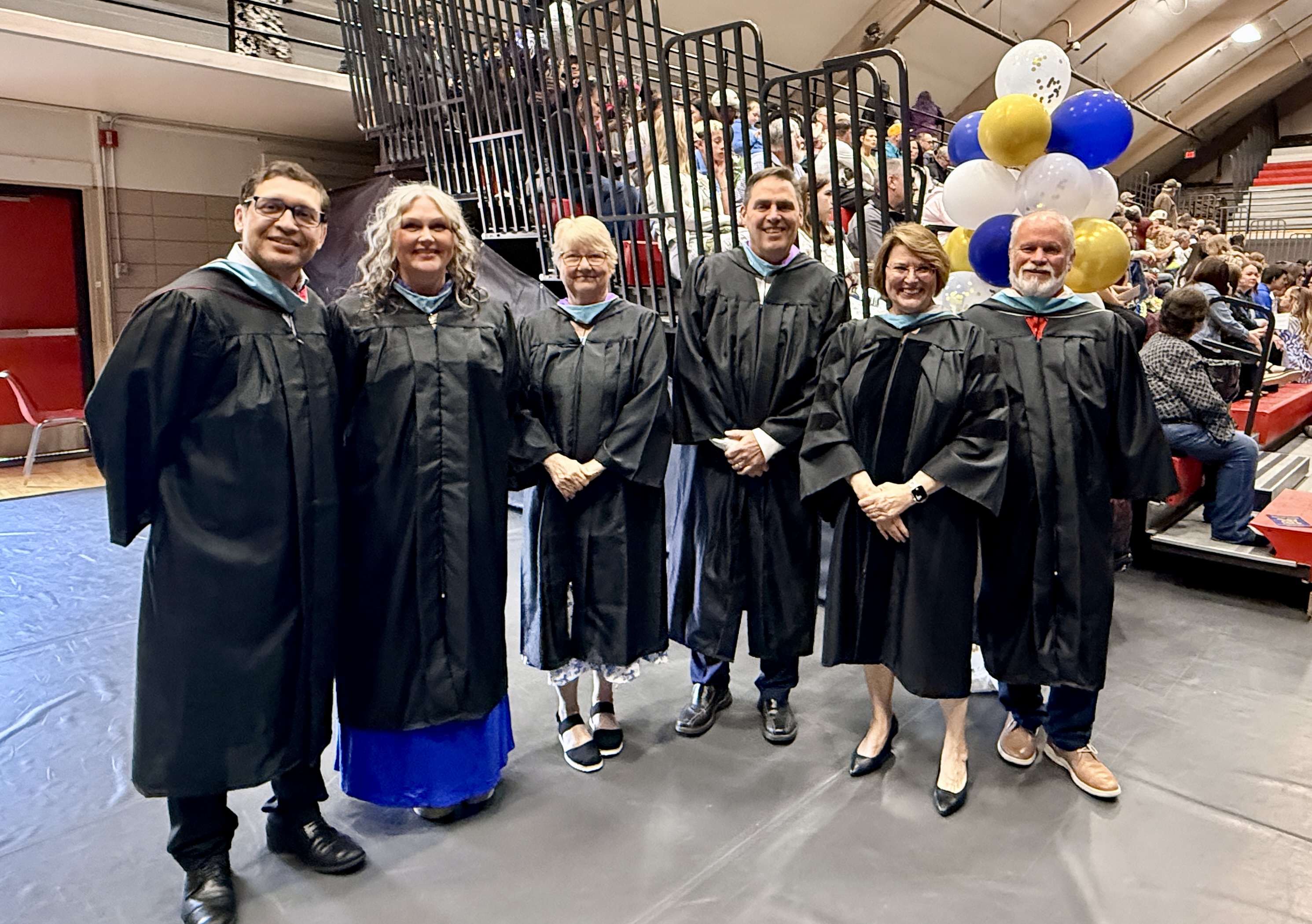 Six people in caps and gowns pose for a photo in an indoor auditorium with stairs, balloons, and a crowd.