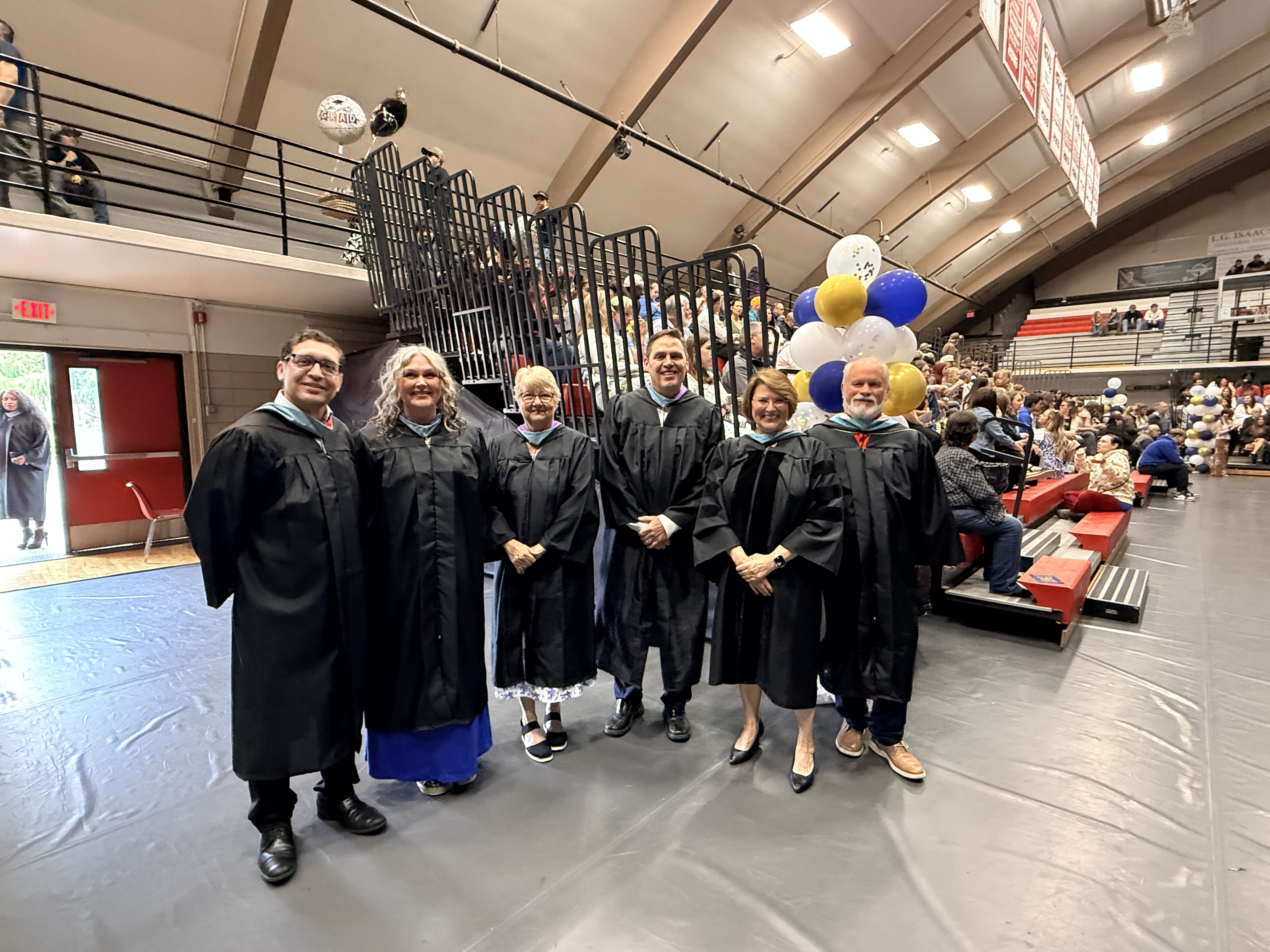 Several people in black gowns stand together in a large hall with red bleachers and balloons.