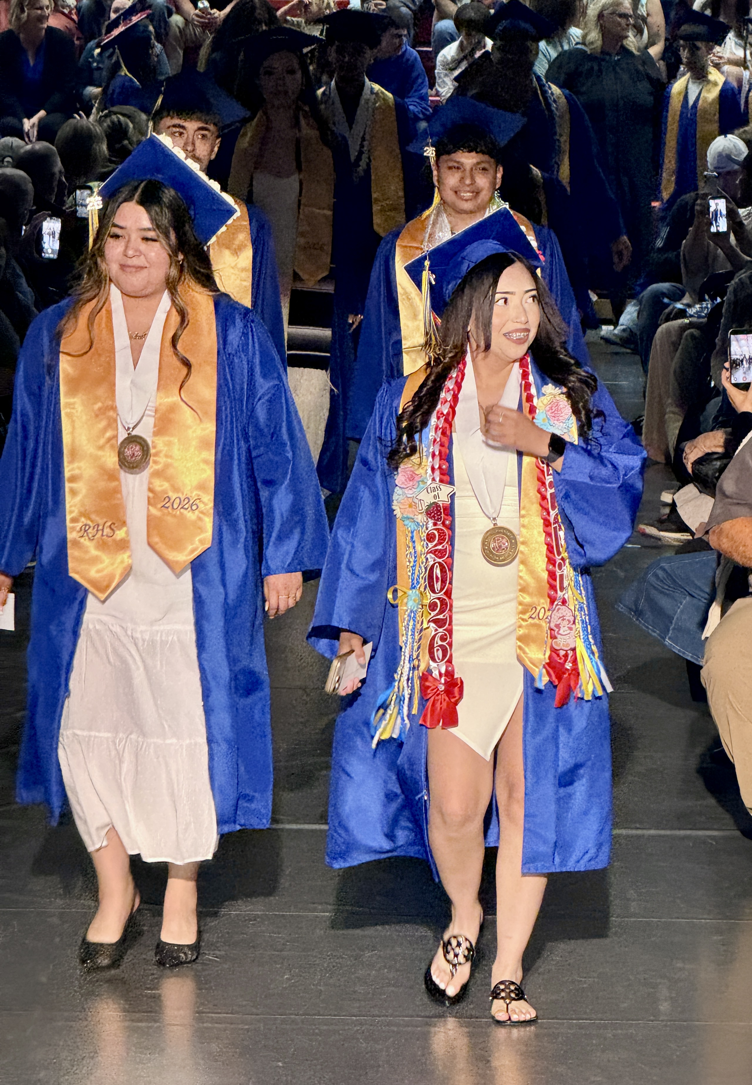 Graduates in blue gowns walk through a stadium. One wears a white dress and the other a tan dress.