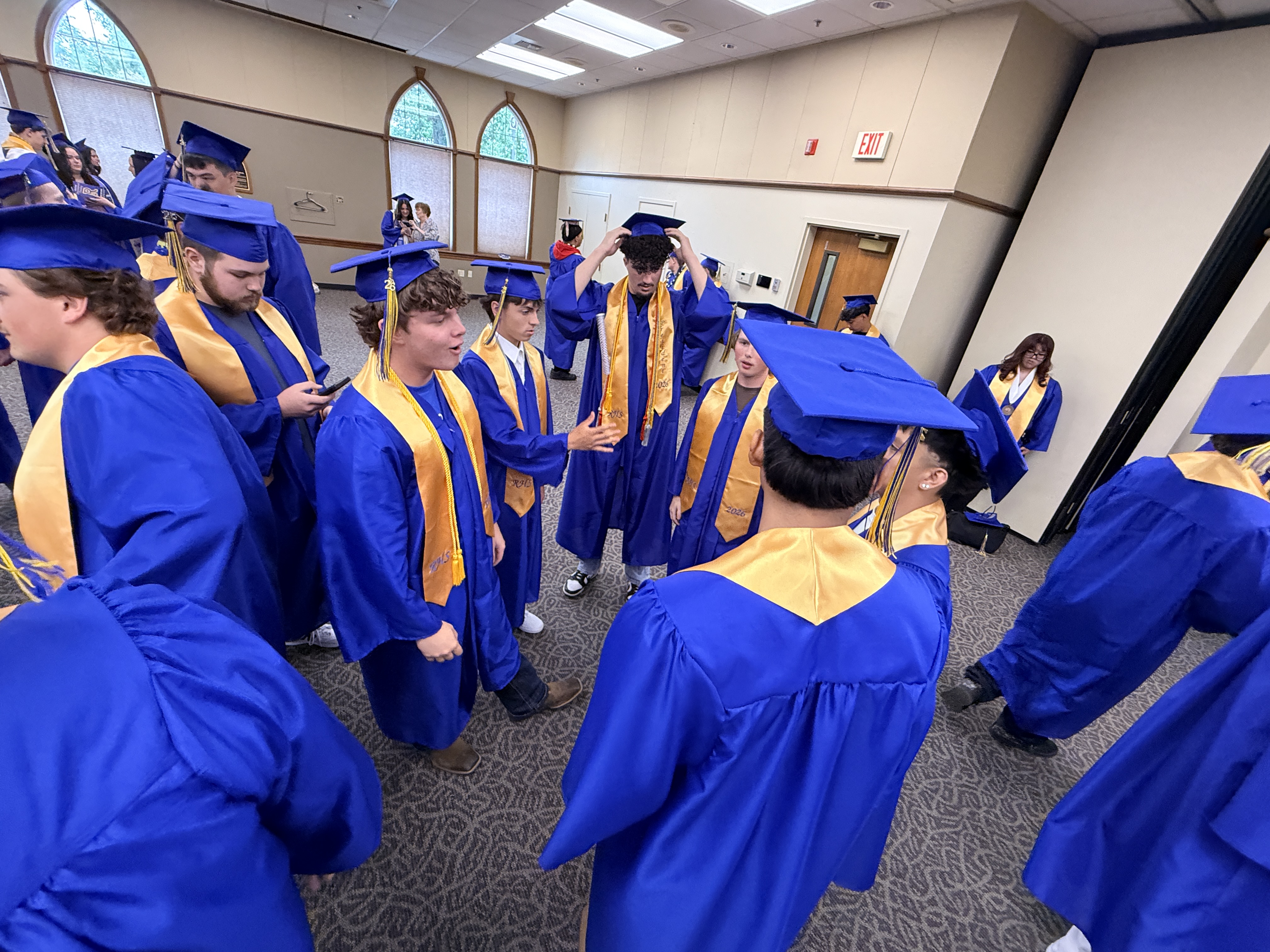 Group of students in blue and gold caps and gowns in a room. A woman in the background is observing.