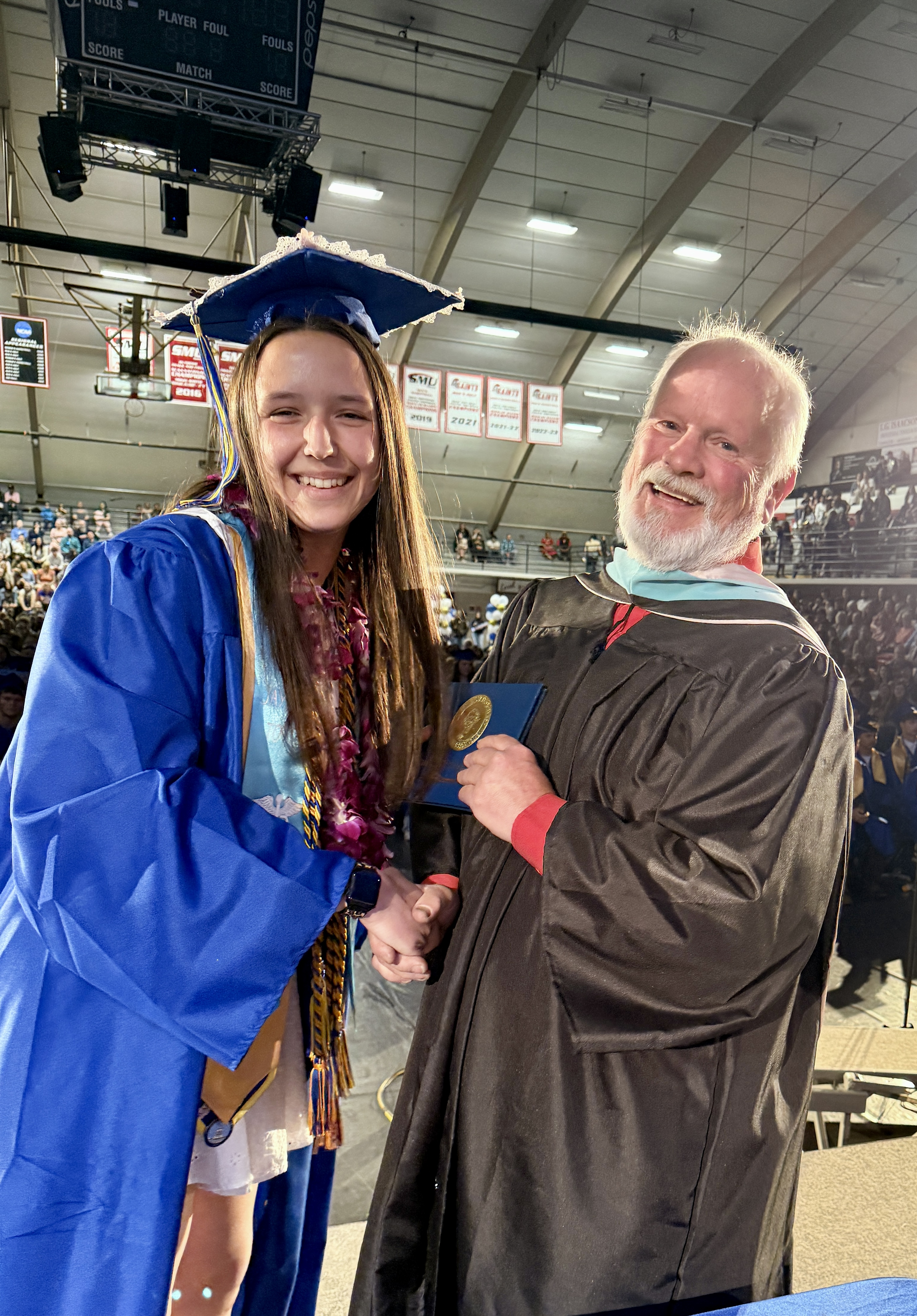 A woman in a blue graduation gown shakes hands with a man in a black gown.