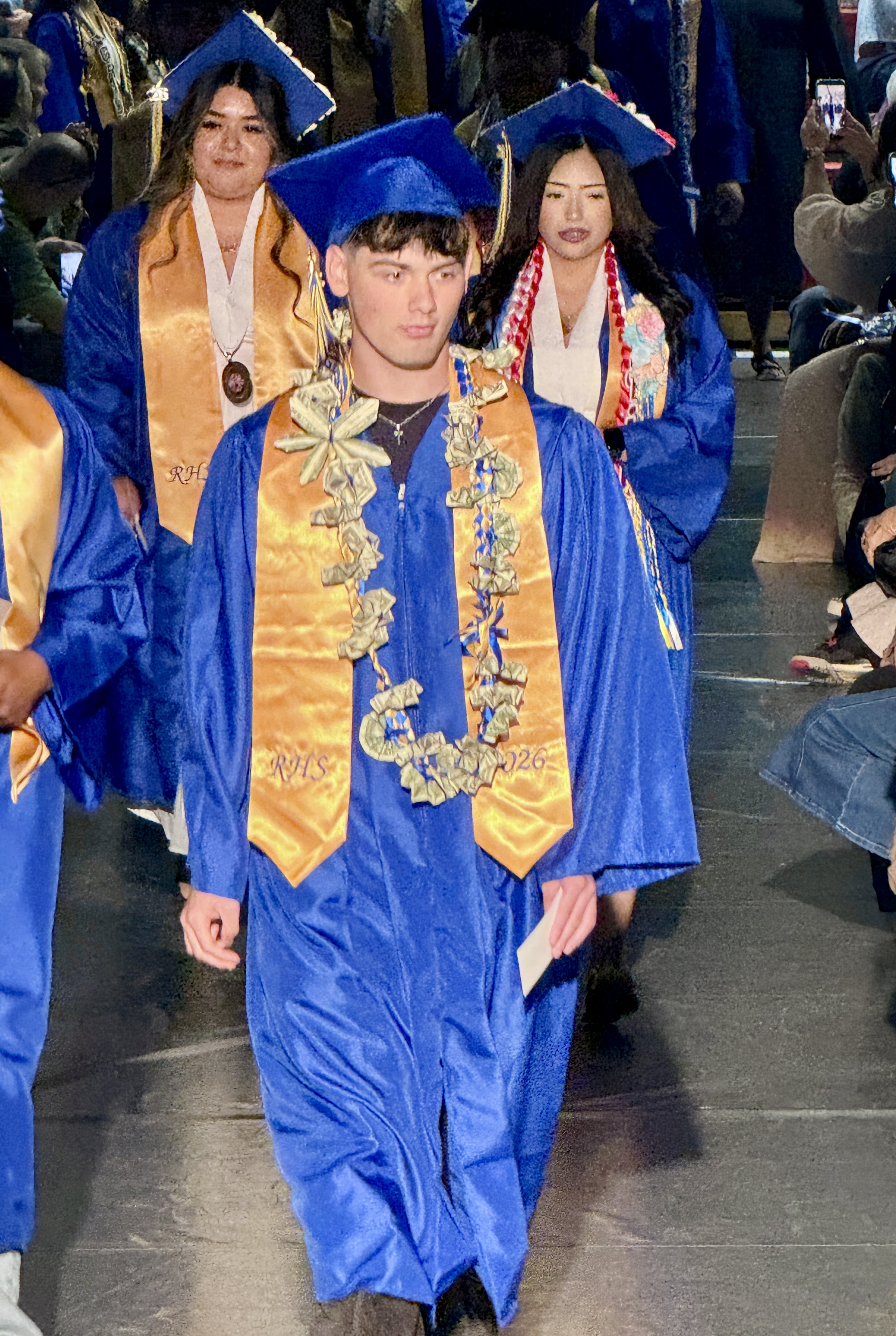 Graduates in blue caps and gowns walk in a ceremony. They wear leis, medals, and hold papers. Audience members watch.