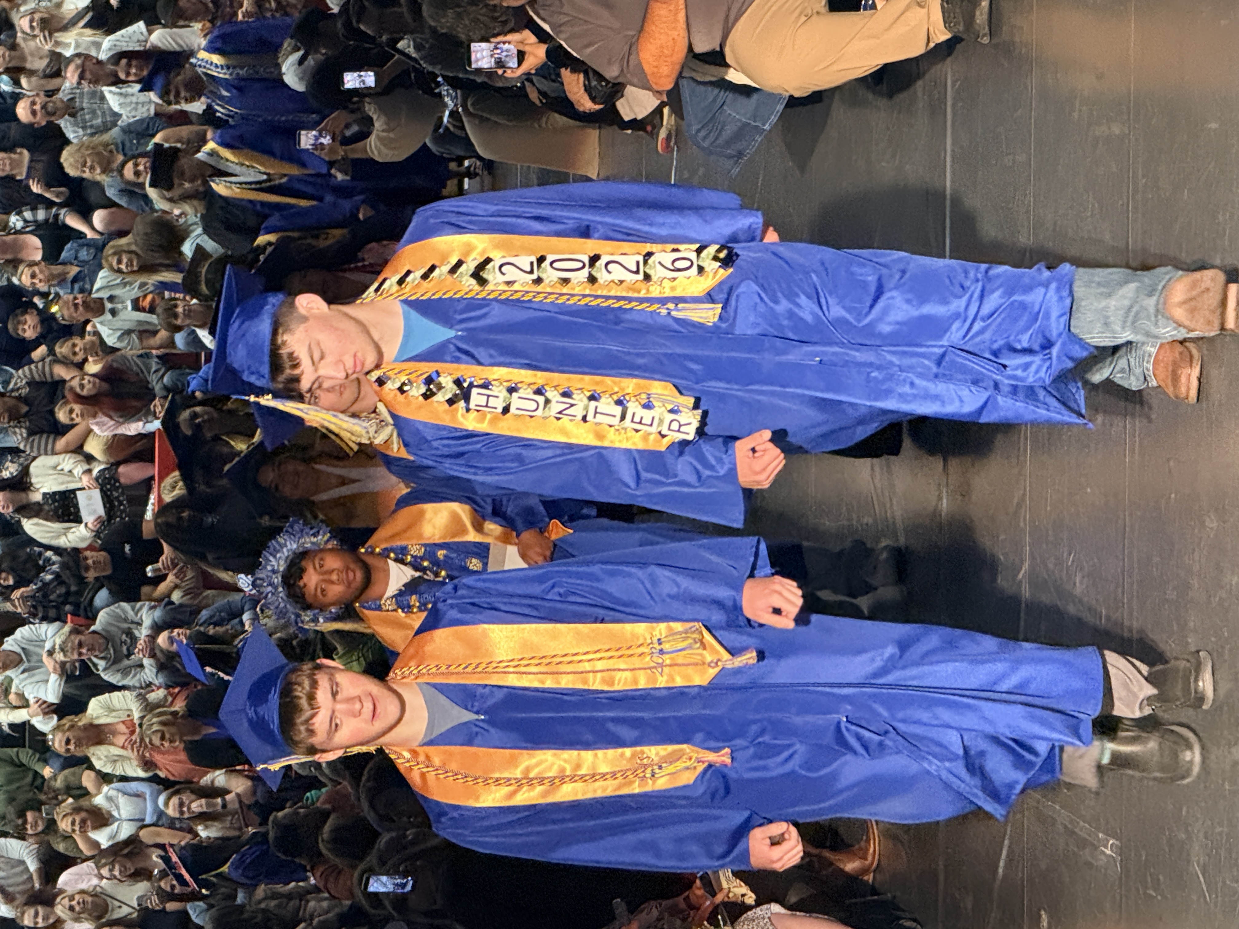 Two young men in blue and gold gowns walk on a stage in front of a seated audience.