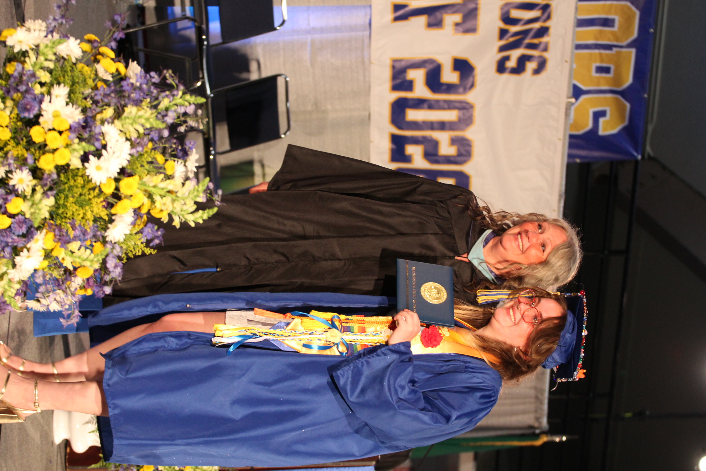 A woman in a black gown presents an award to another in a blue gown at a graduation ceremony. Behind them, a banner reads "Graduation 2023."