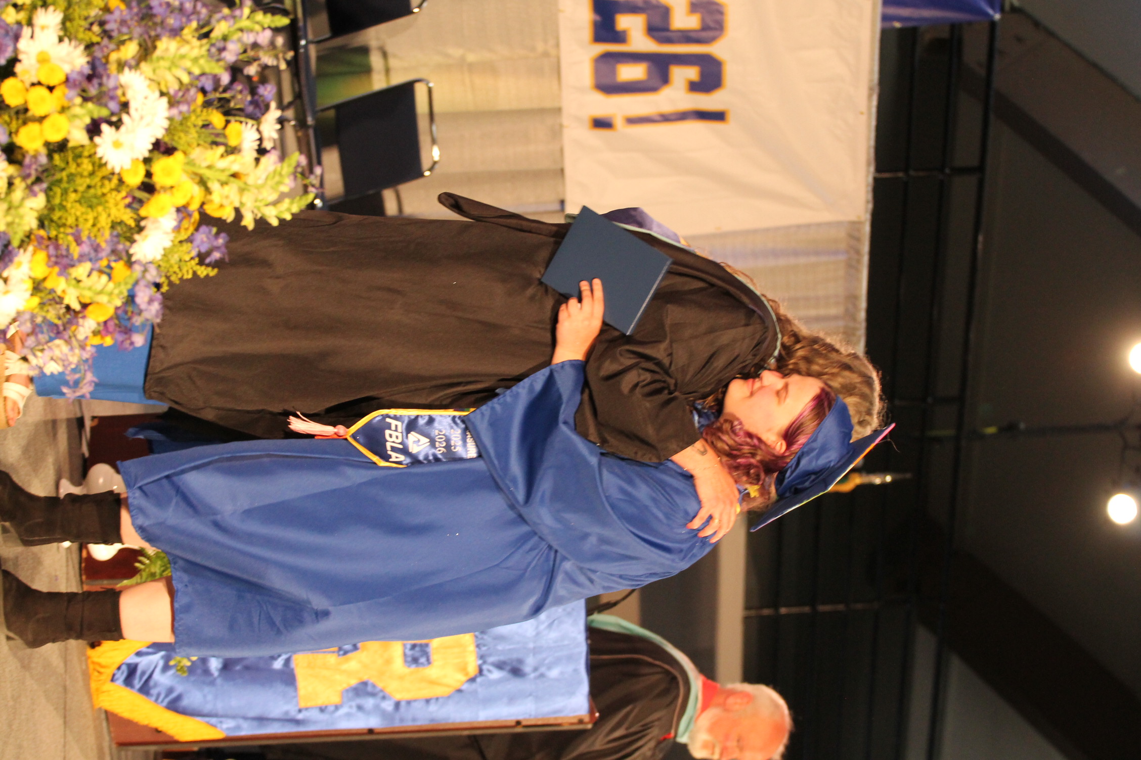 Two women in graduation gowns hug each other, one holding a diploma, on a stage with flowers.