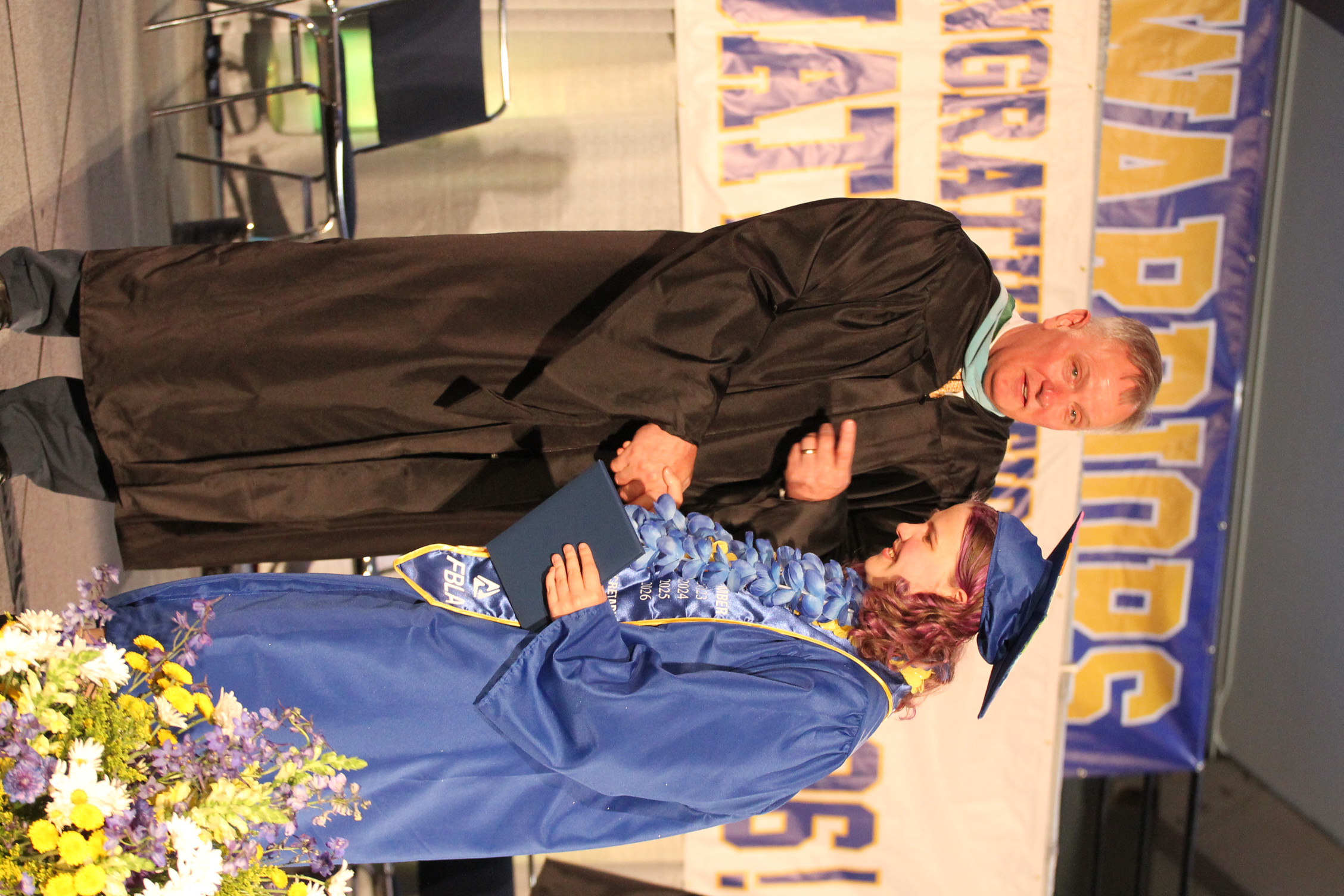 Man and woman in graduation gowns on a stage. He hands her a diploma. A banner reads "Congratulations!"