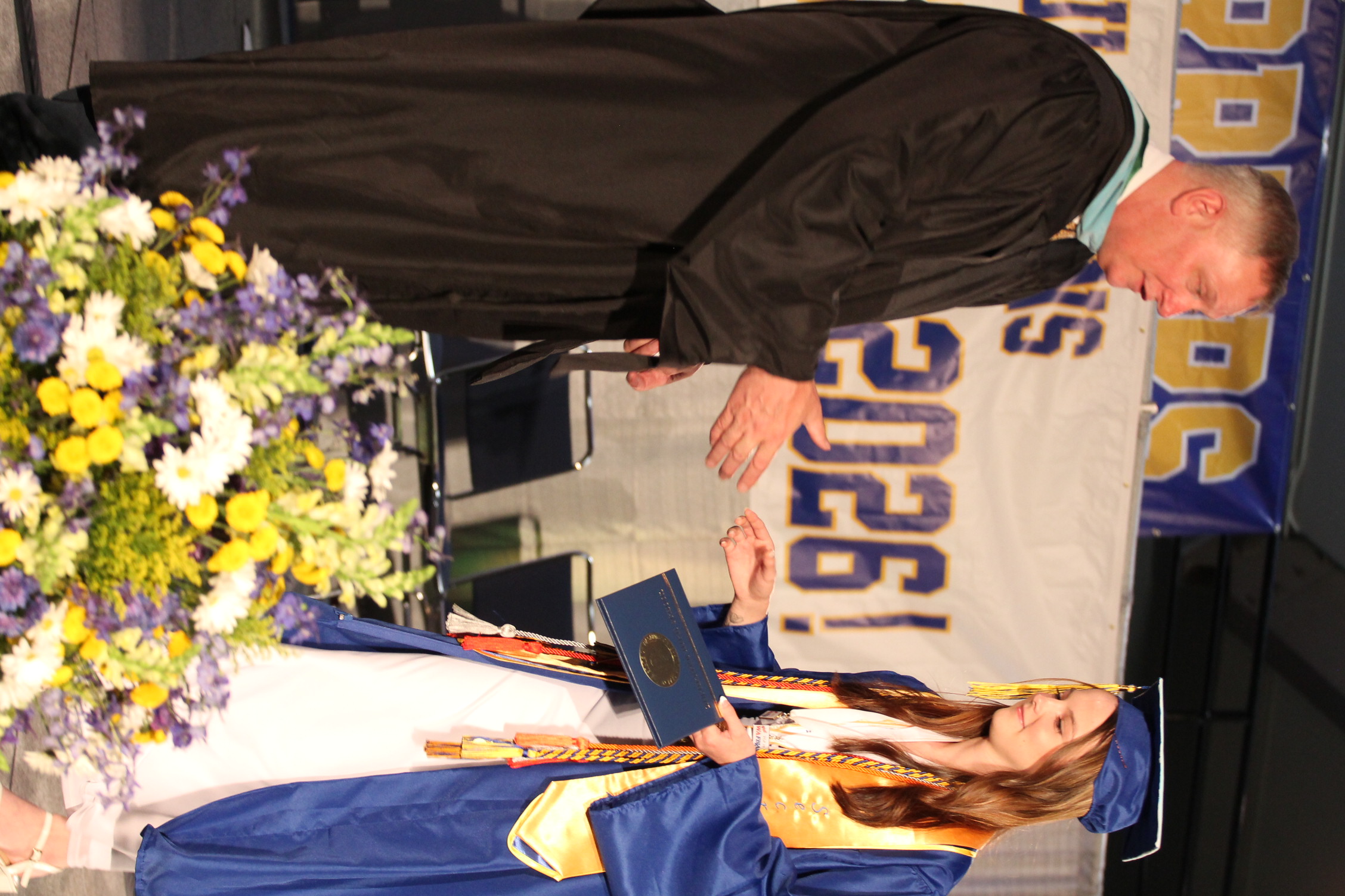 A man and a woman stand on stage. The man in a black gown hands the woman a diploma. She wears a blue and white graduation gown and cap.