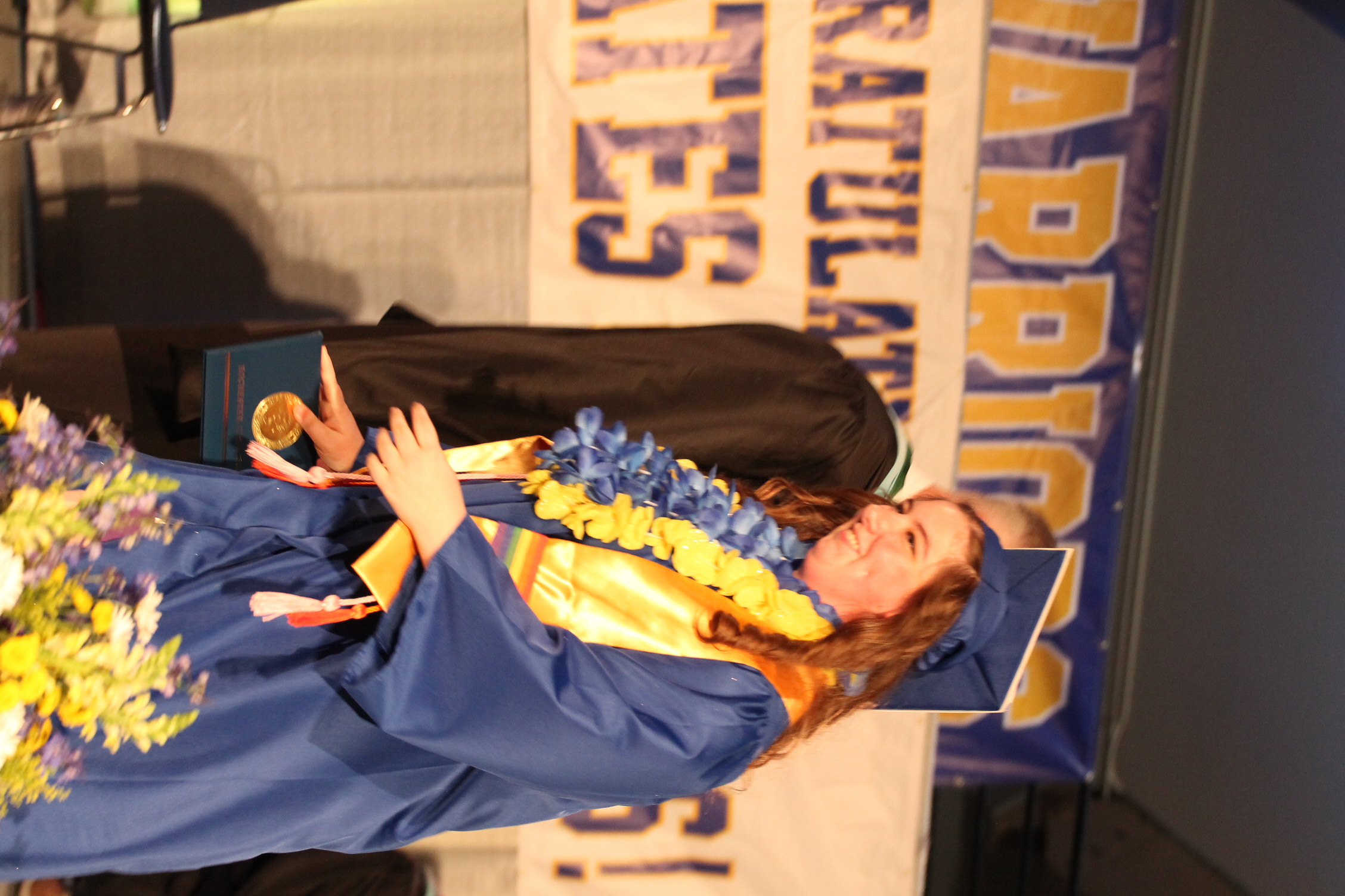 A person in a blue graduation gown stands on a stage, holding a diploma, and smiles. A banner with "Warriors" and "Congratulations" hangs behind them.