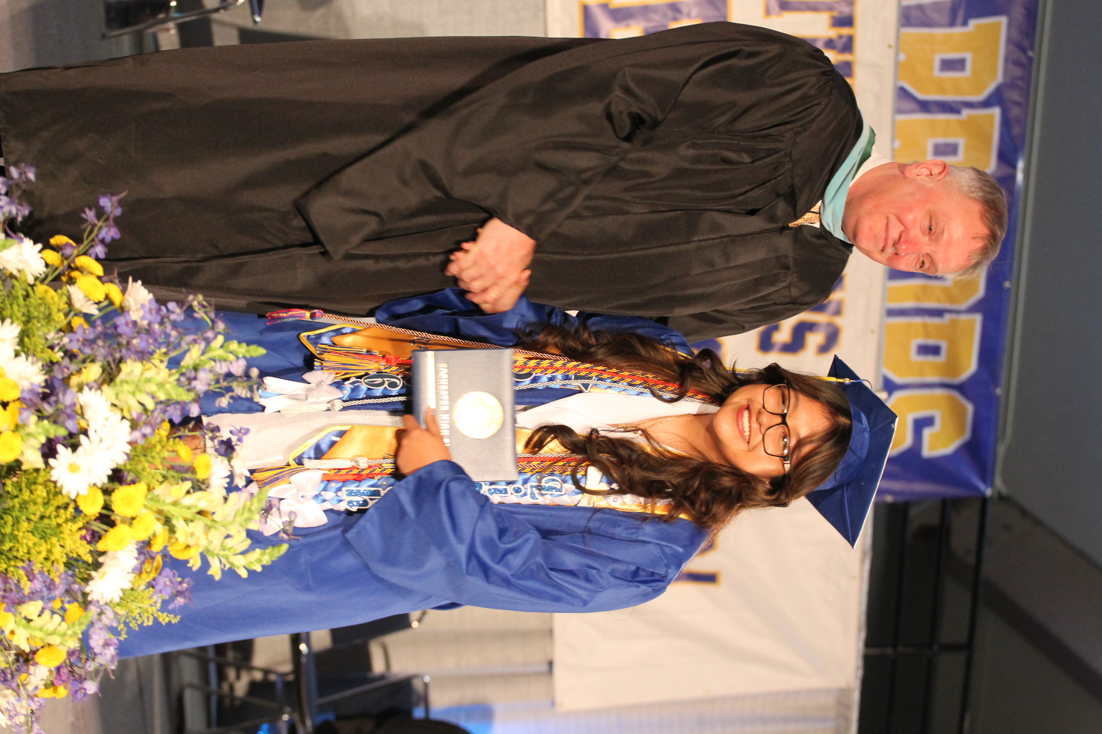 A man and a woman in caps and gowns stand on a stage with a bouquet, shaking hands. A banner is in the background.