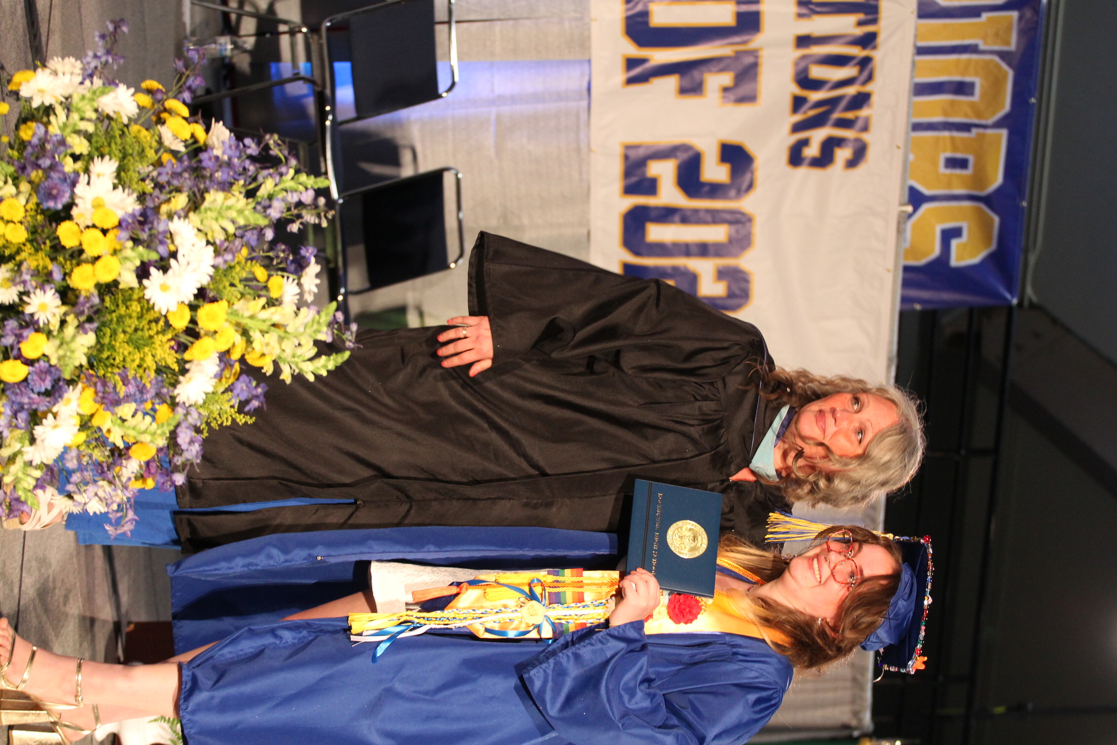 Two women in graduation robes and caps stand on a stage. One holds a diploma. Behind them, a banner reads "Congratulations 2023."