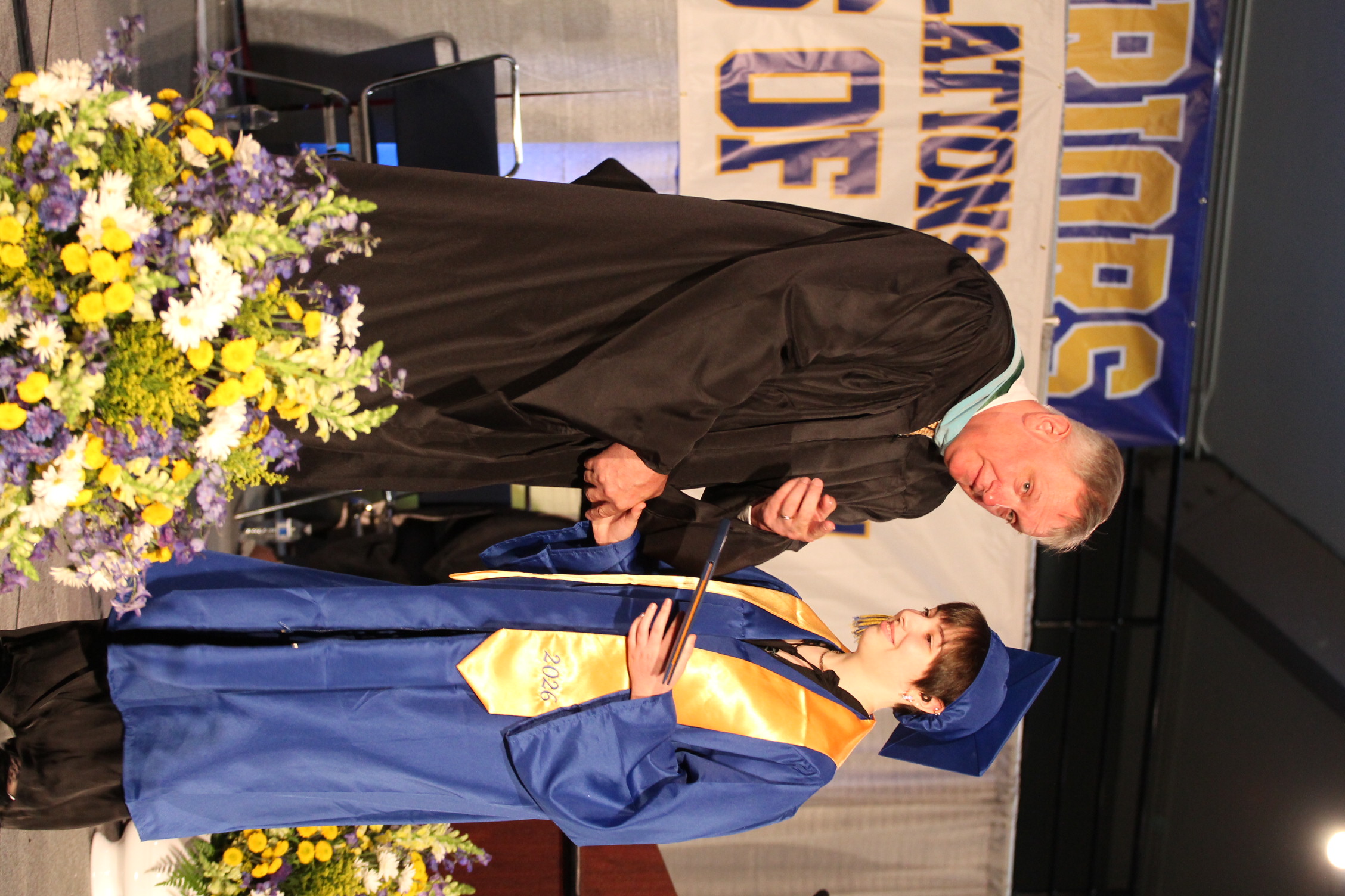 A man in a black robe hands a diploma to a student in a blue graduation gown.