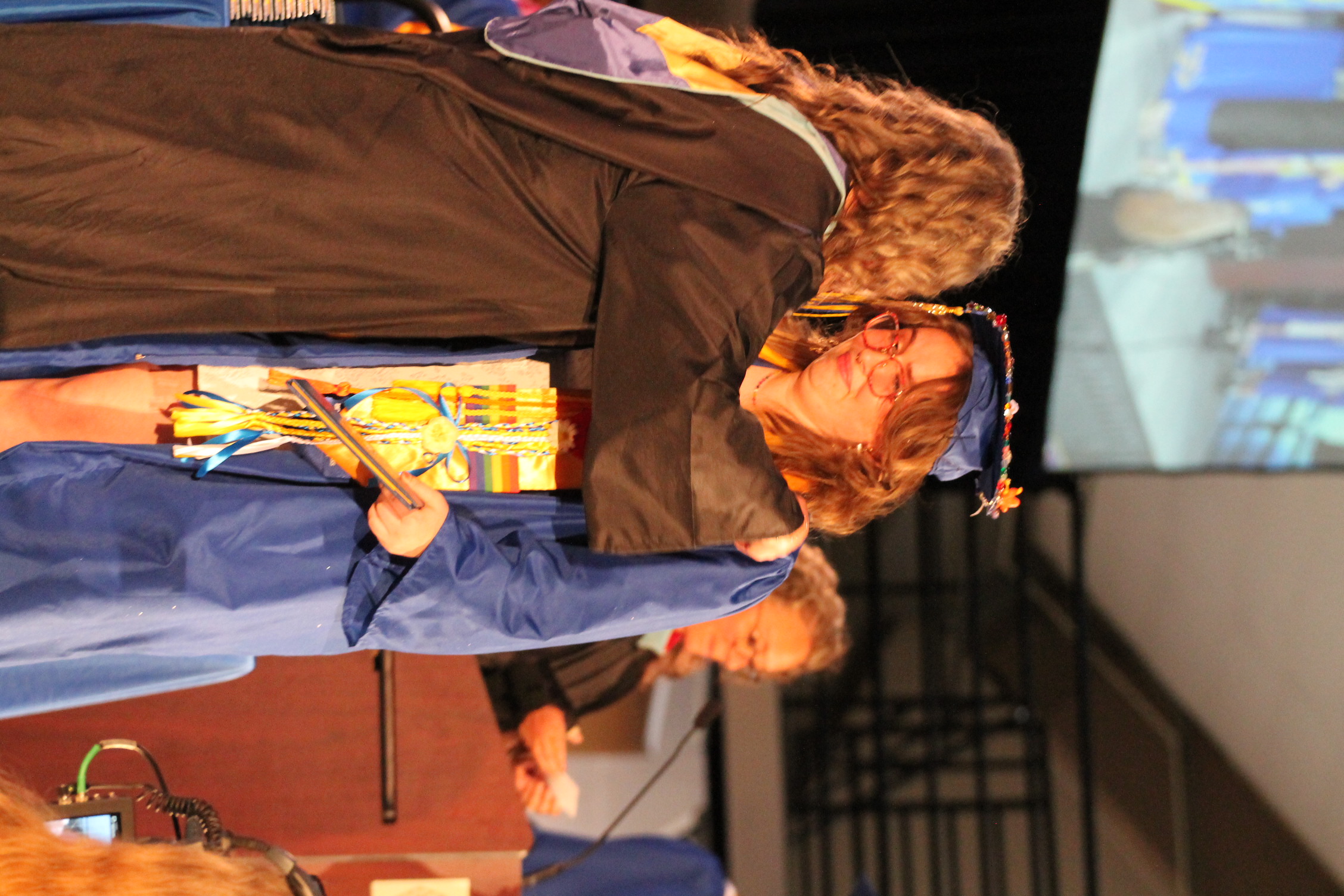 Woman in cap and gown receiving congratulations from another woman in a similar outfit on stage.