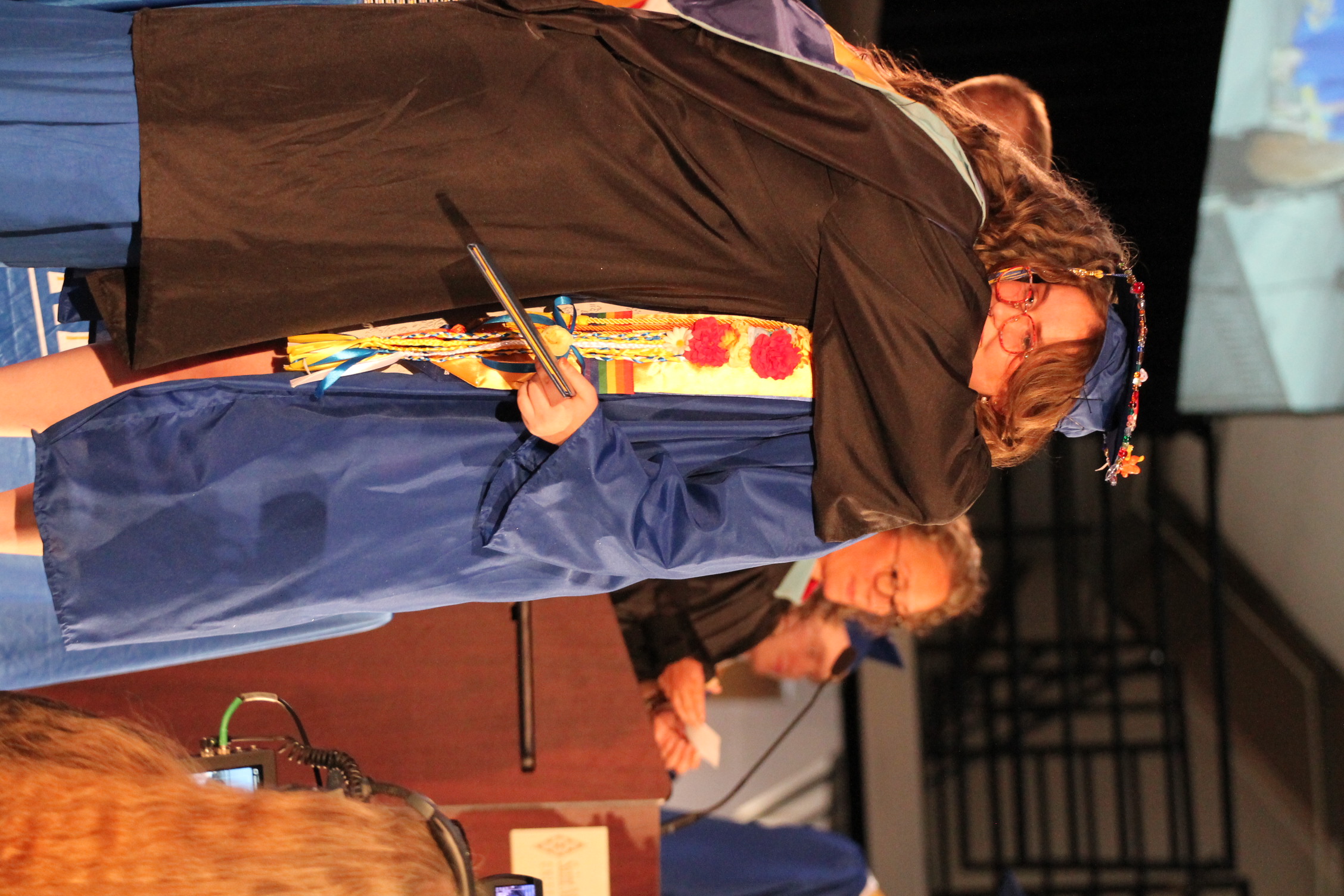 Two women in blue graduation gowns hug, one holding a diploma, on a stage with a podium.