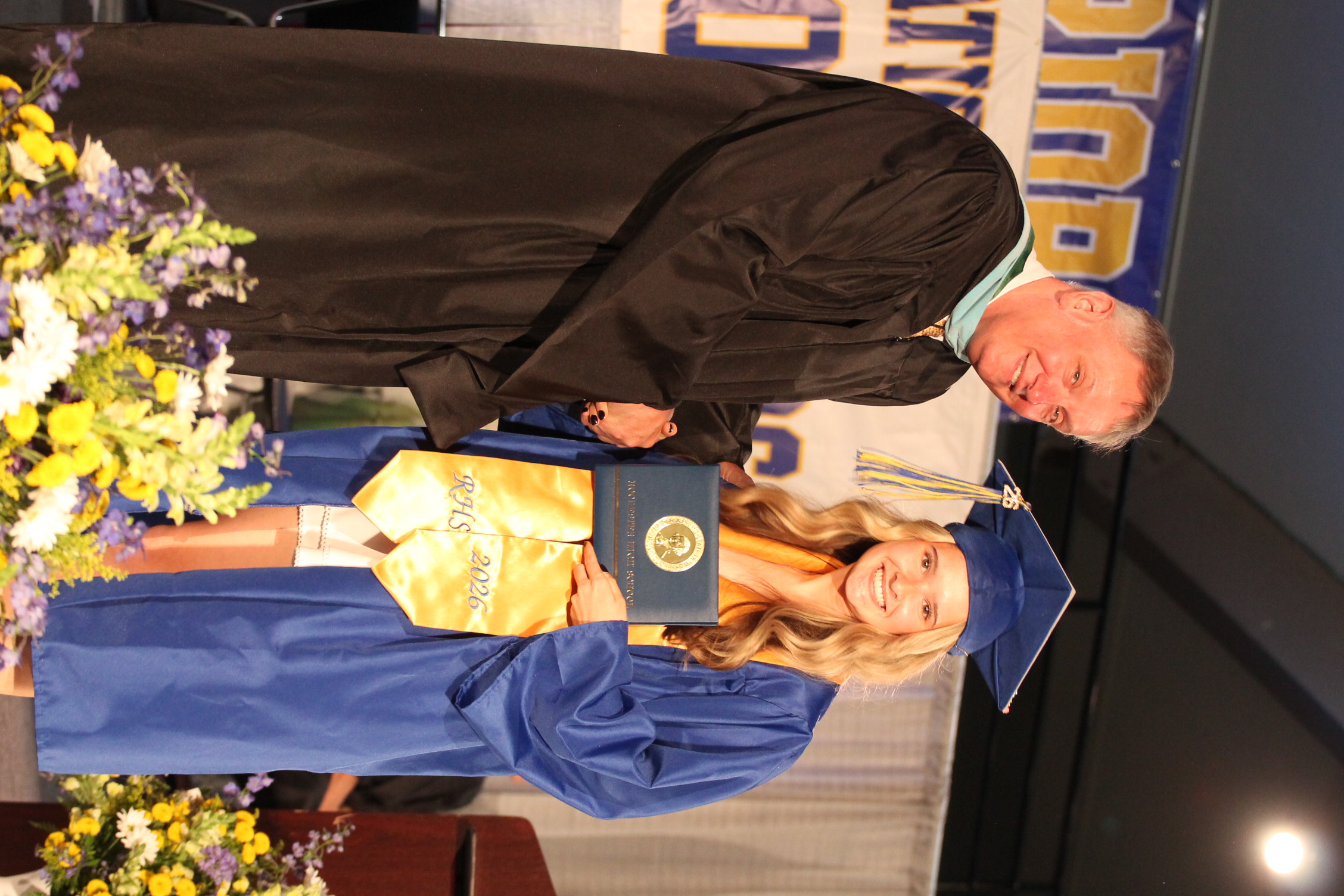 A male wearing a black gown hands a framed certificate to a young woman in a blue gown and cap.