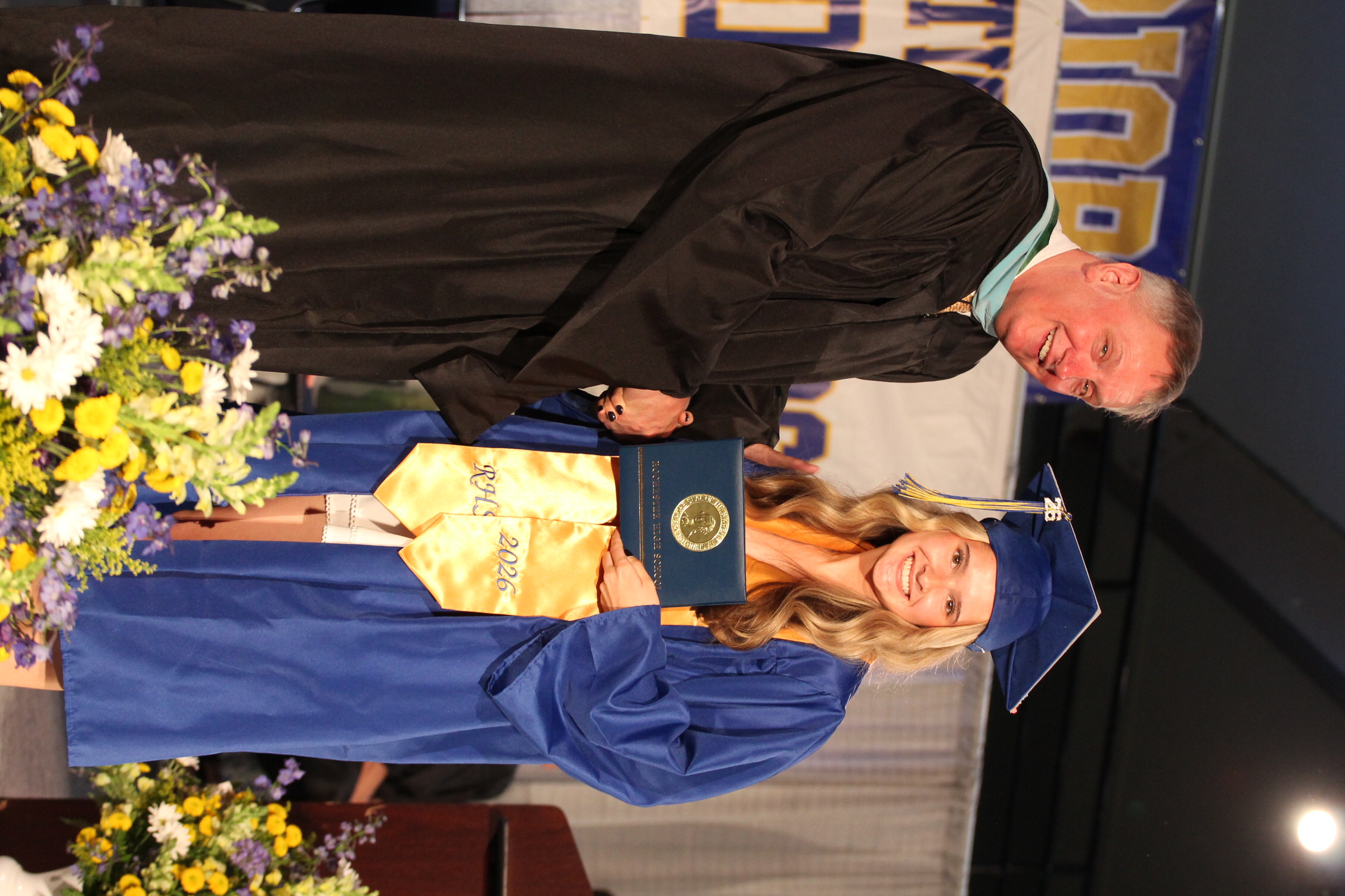 A smiling man in a black robe shakes hands with a woman in a blue gown and cap holding a framed certificate.