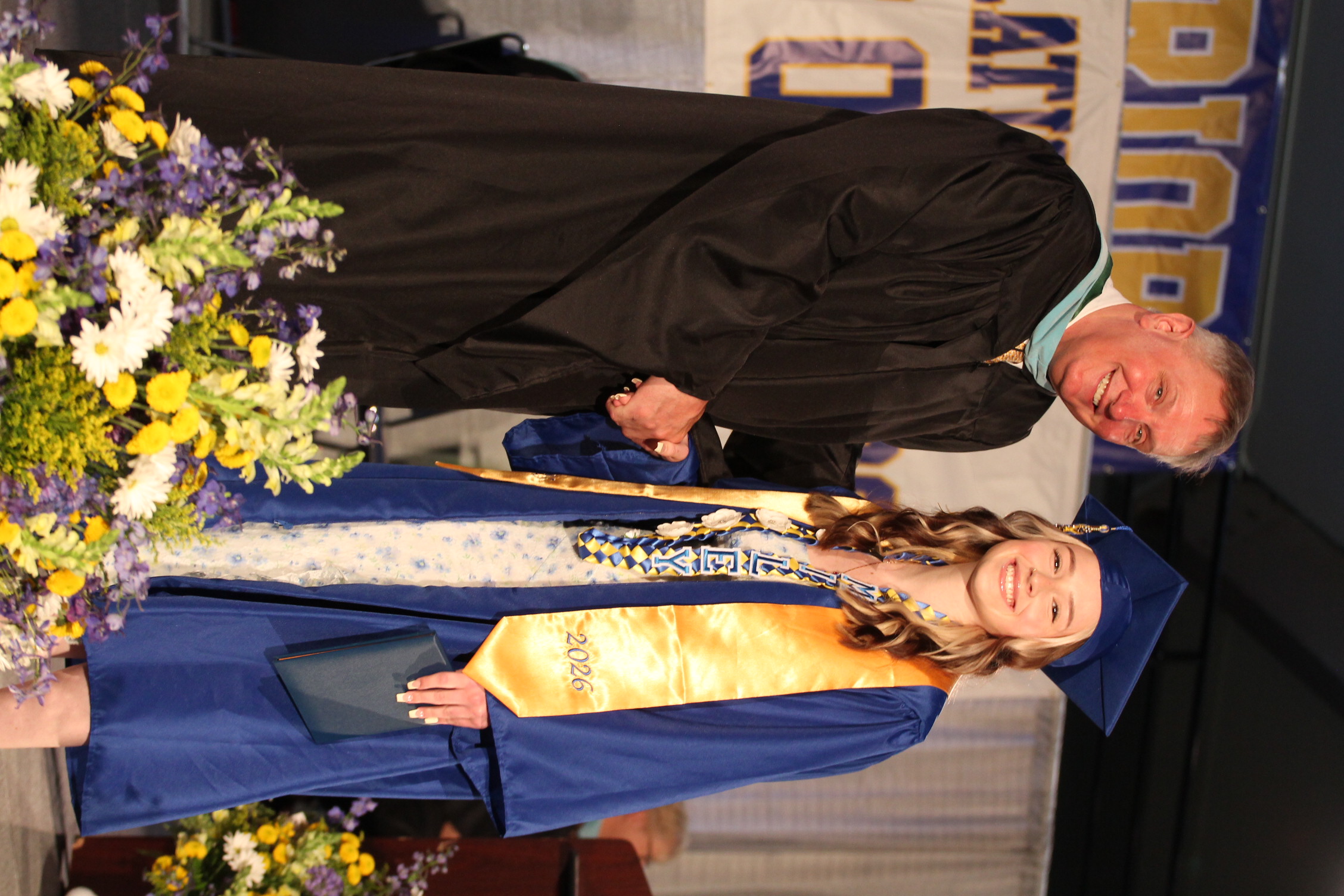 A man and woman stand together on a stage, the man wearing a black gown and the woman in a blue gown.