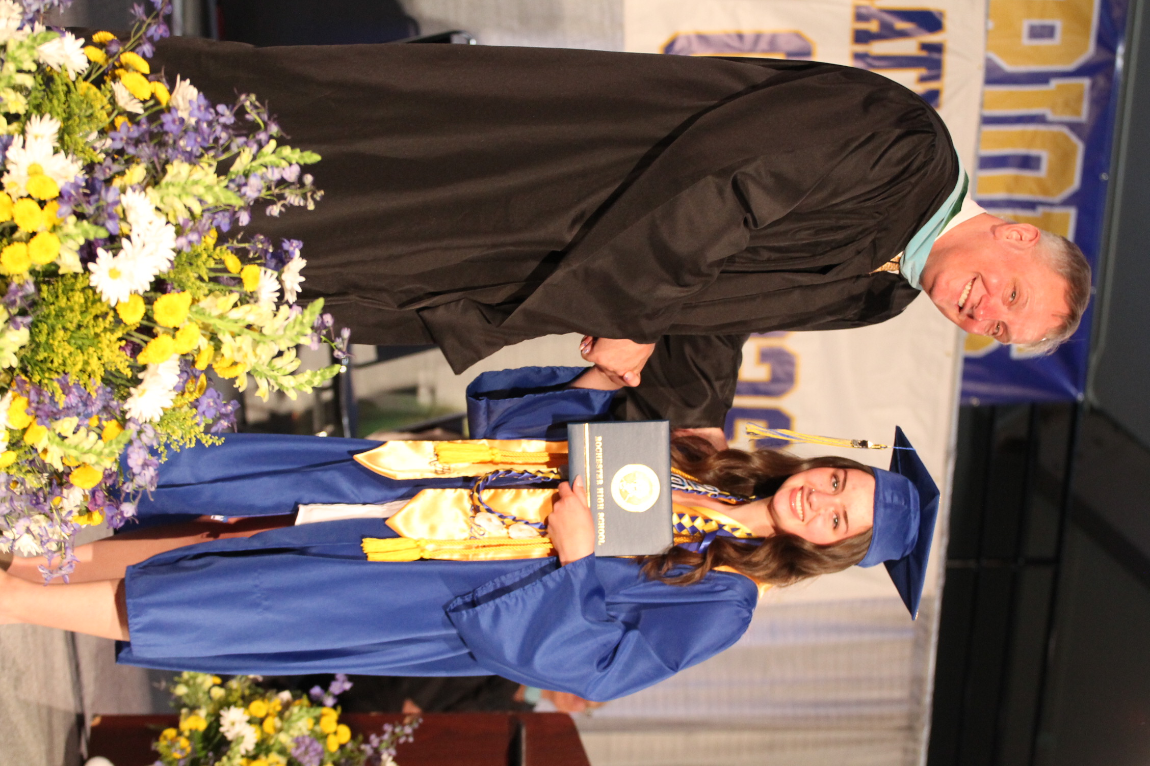 A man in a black gown shakes hands with a woman in a blue gown at a graduation ceremony.