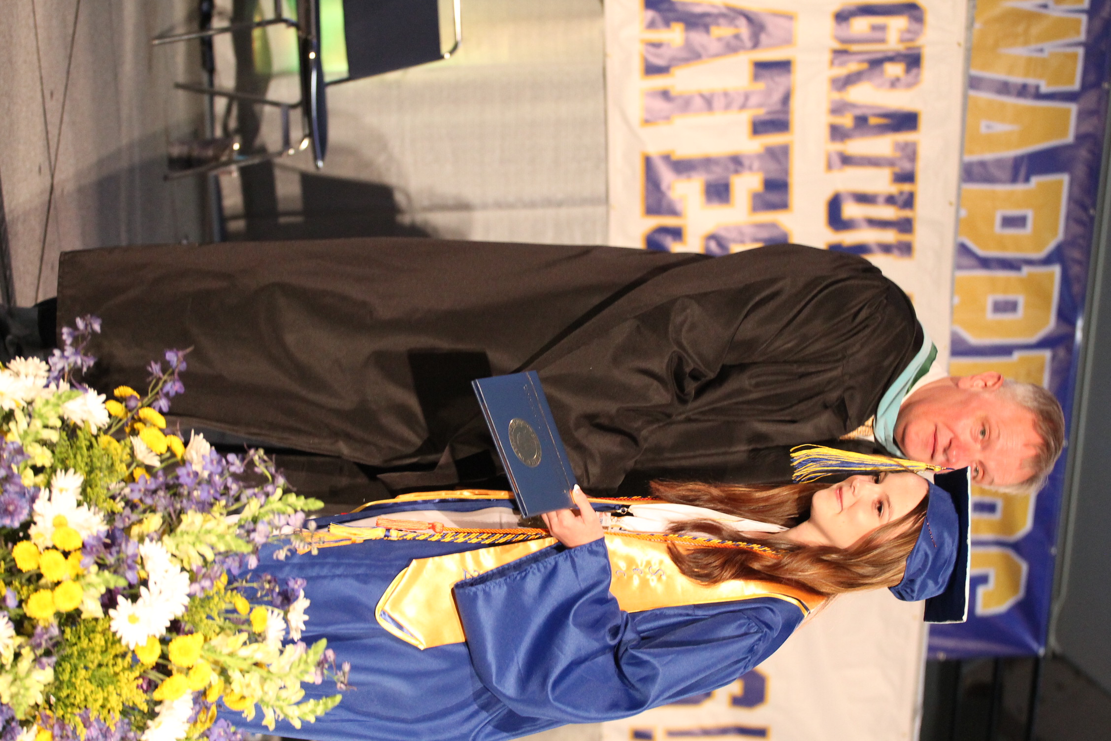 A man in a black gown hands a diploma to a woman in a blue gown. Behind them is a stage with a banner.