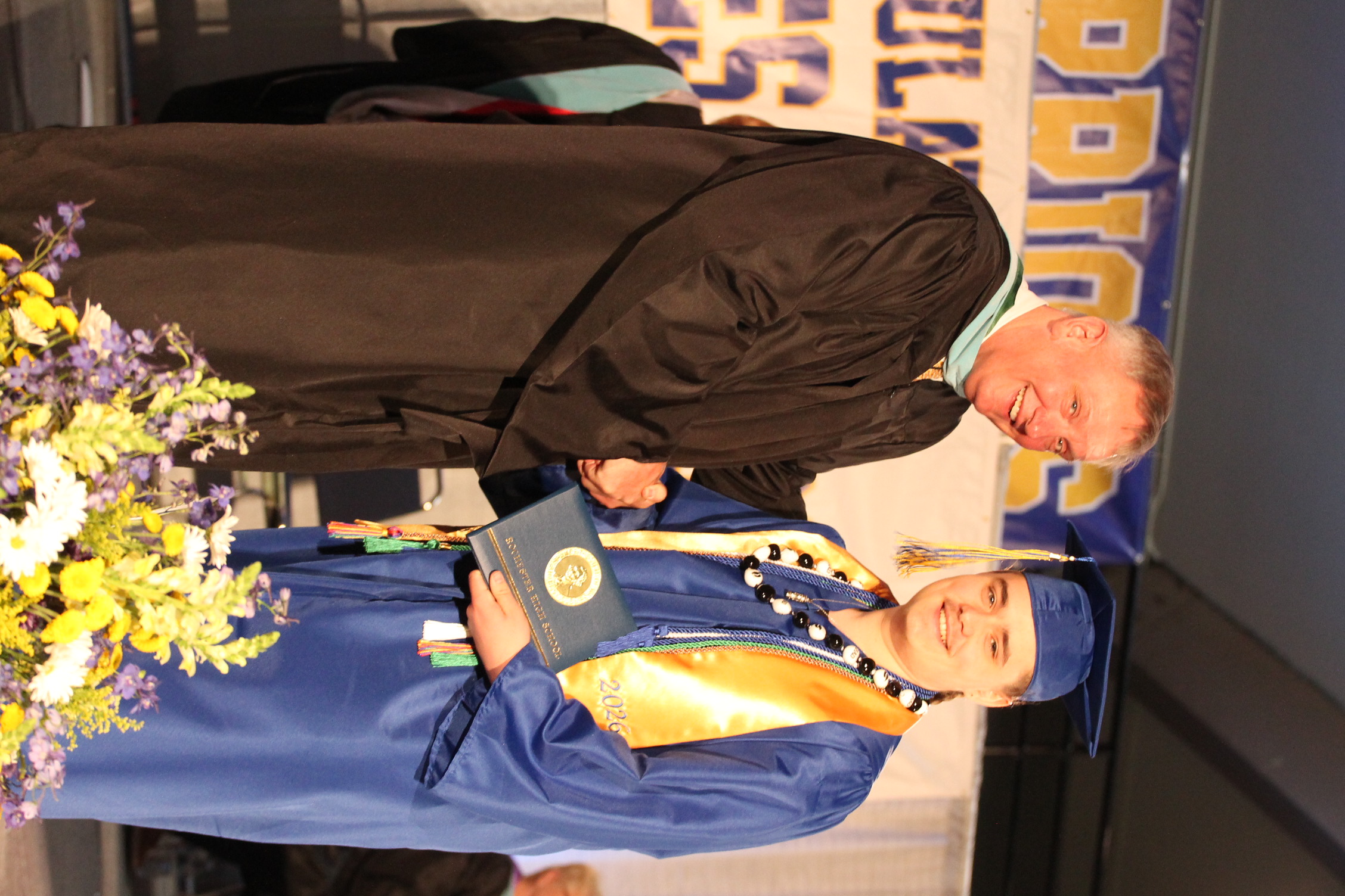 A man in a black gown hands a diploma to a man in a blue gown. They shake hands.