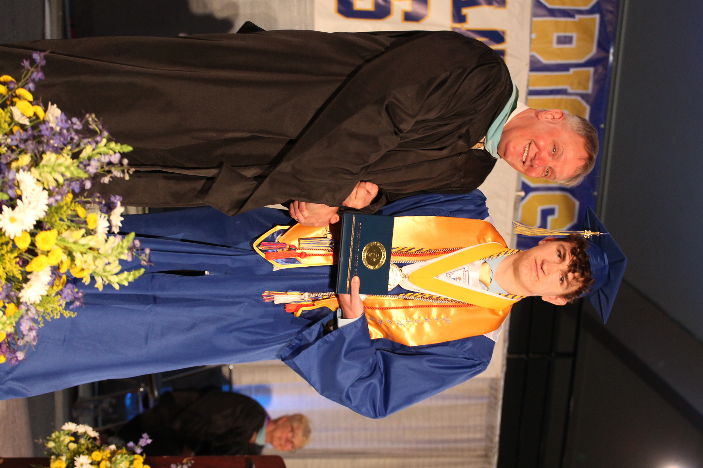 A man in a cap and gown receives an award from another man in a black gown. Behind them is a banner, flowers, and another individual seated.