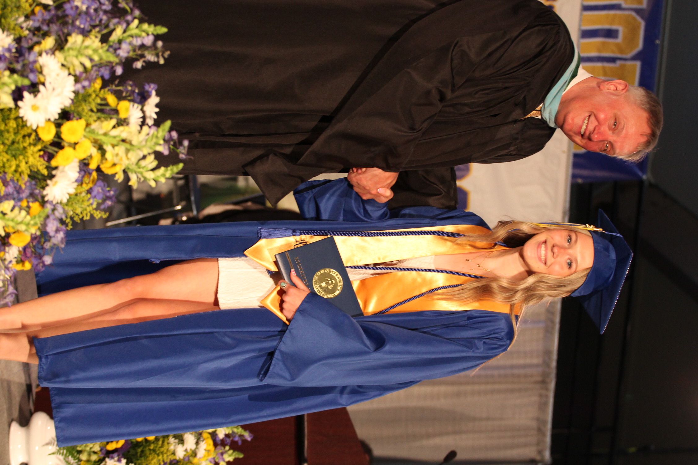 A smiling man in a black gown shakes hands with a woman in a blue gown at a graduation.