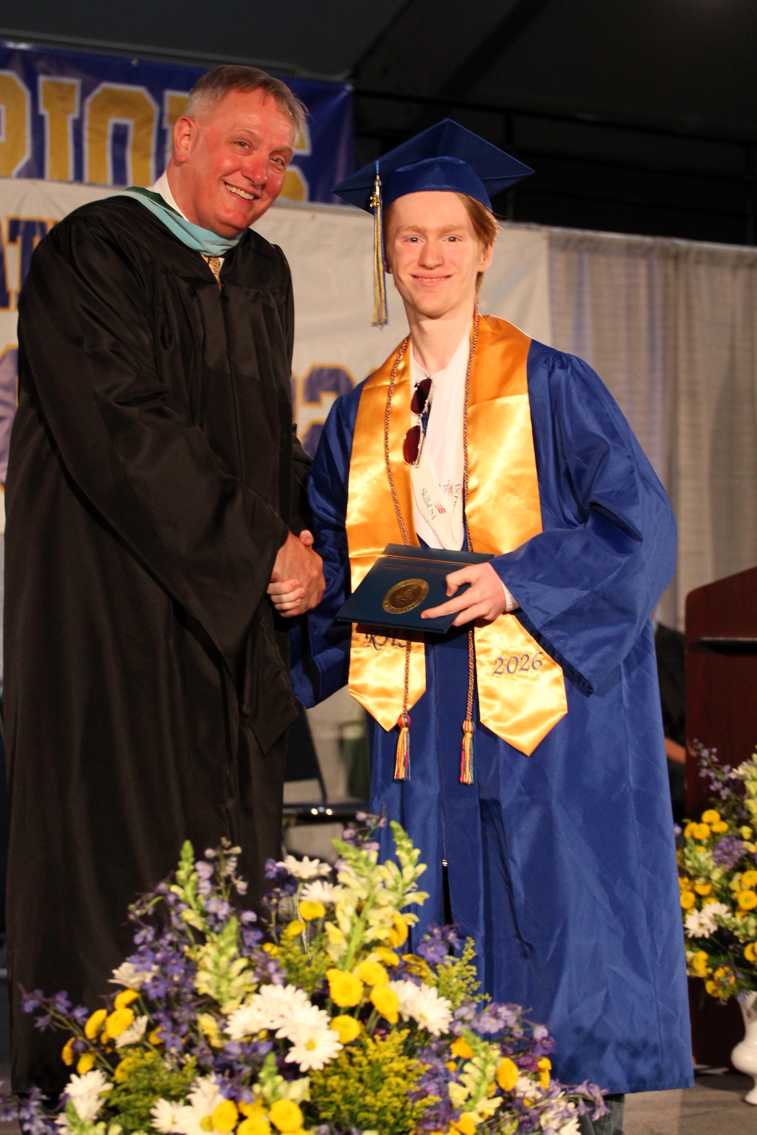 A man in a black gown hands a diploma to a student wearing a blue gown and cap, both smiling. Flowers are in the foreground.