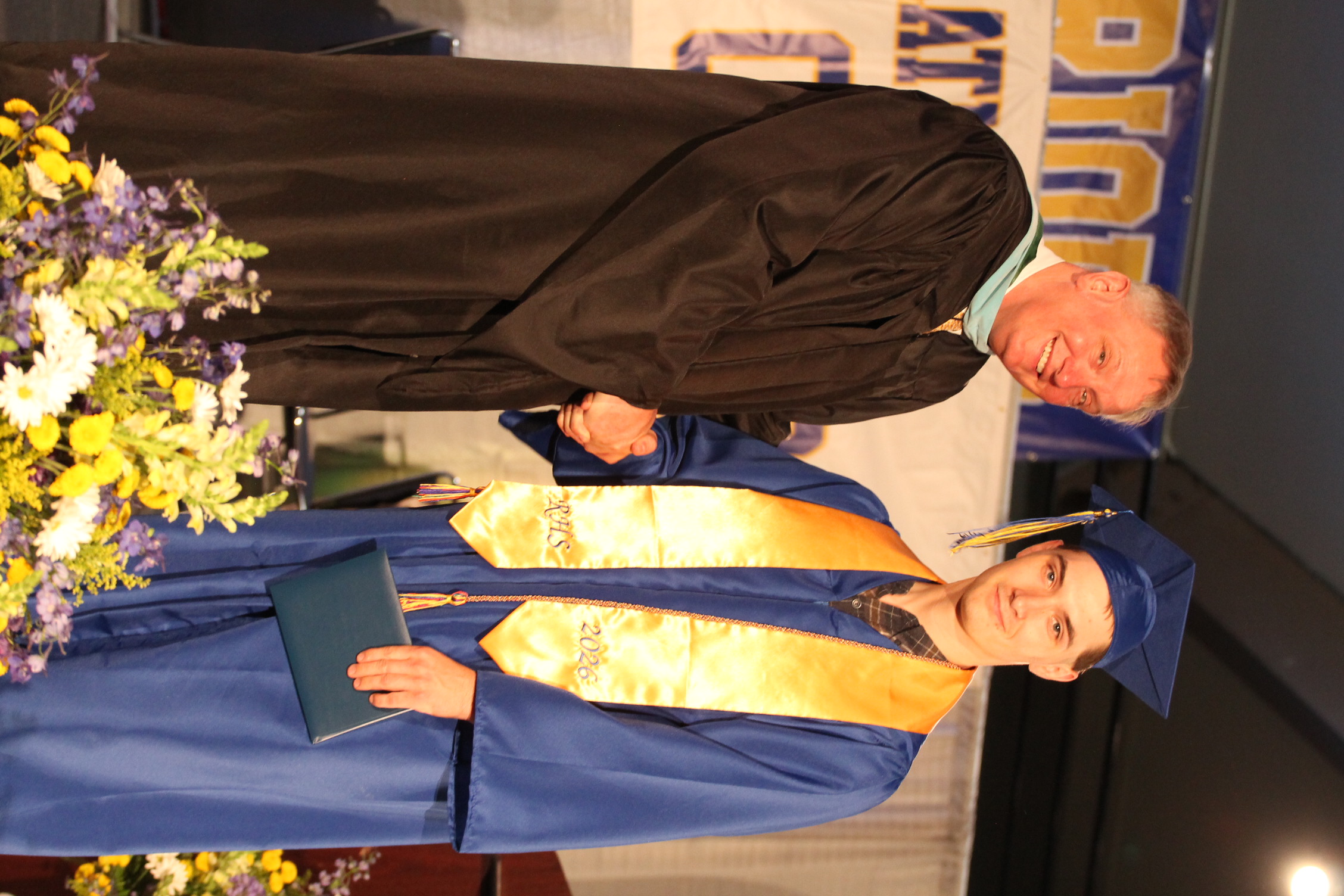Man in academic regalia, shaking hands with a graduate, on stage at an event. Flowers in front.