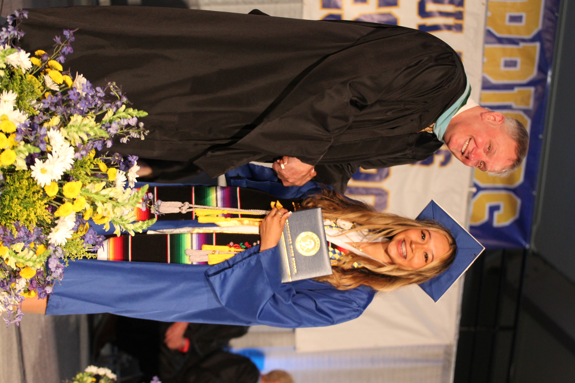 A man in a black robe shakes hands with a woman in a blue graduation gown holding a certificate.