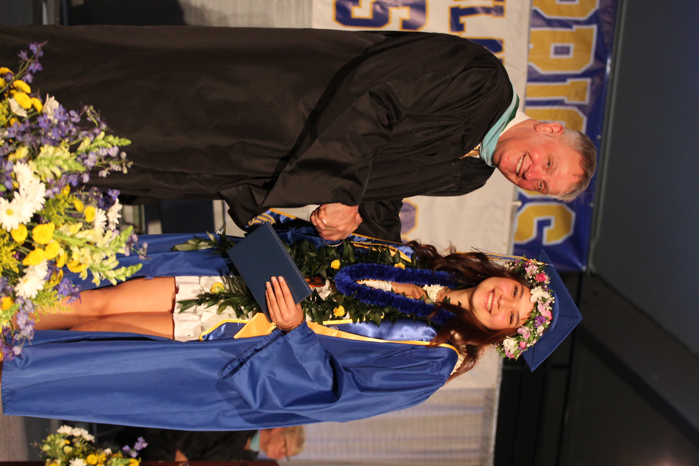 Man and woman in academic gowns. Woman holds diploma and flowers, smiling and shaking hands with man.
