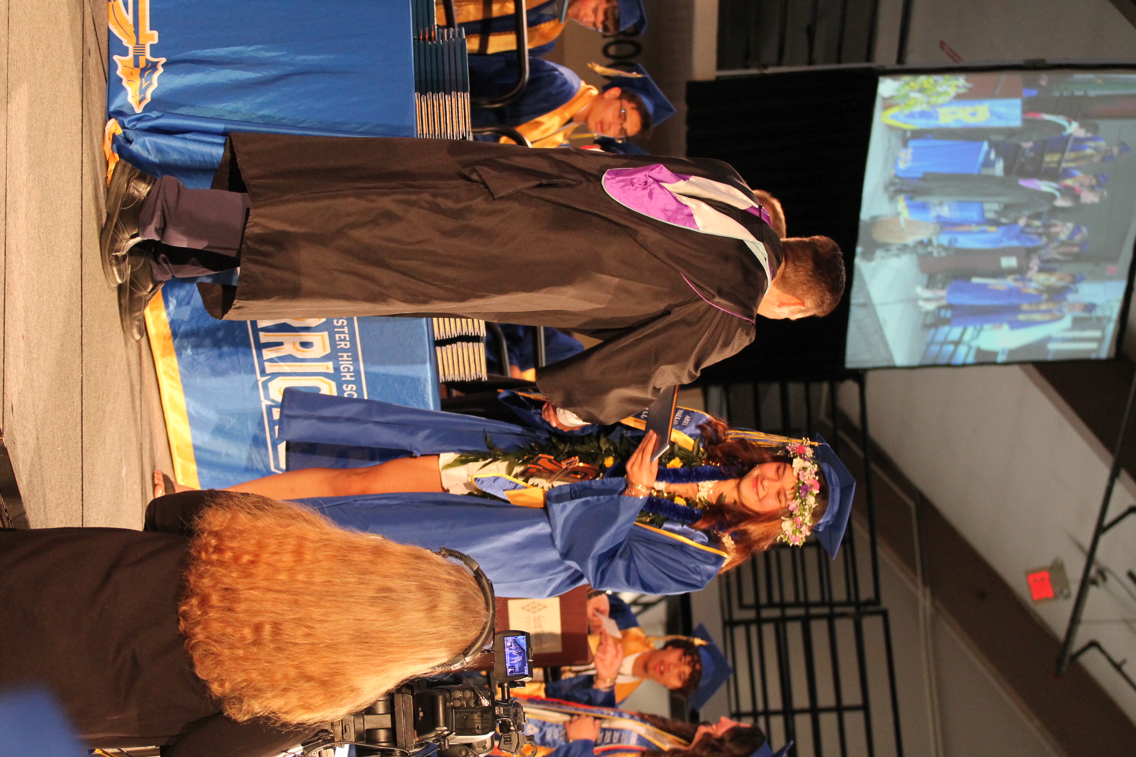 A graduation ceremony with a man and woman on stage, both in gowns. The woman holds a diploma and shakes the man's hand. A screen is visible behind them.