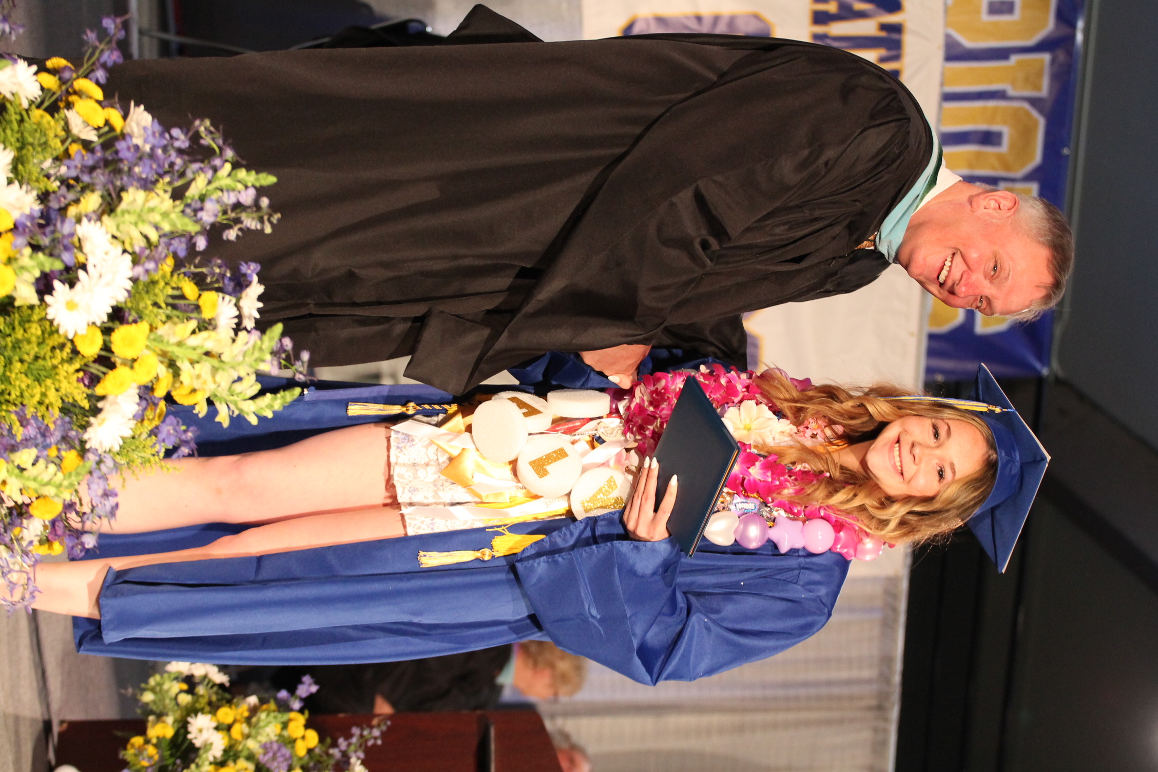 A man in a black gown shakes hands with a woman in a blue gown. They stand on a stage with flowers.
