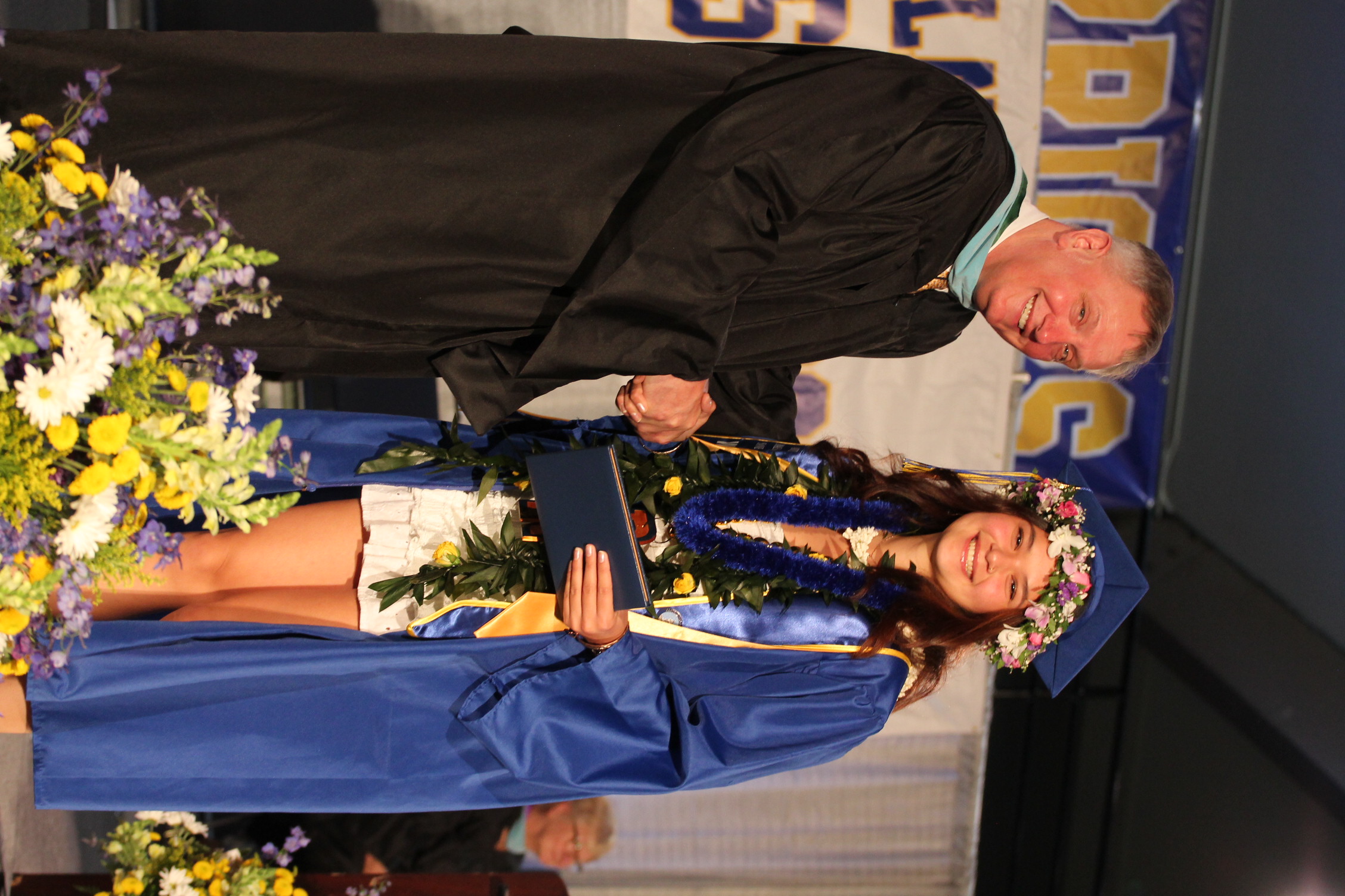 A man and a woman in graduation gowns, with the woman holding a diploma and flowers at the base.