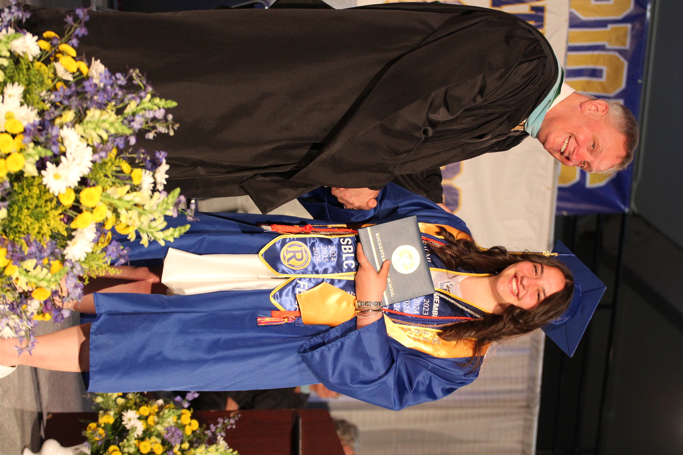 A man in a black gown shakes hands with a woman in a blue gown at a graduation ceremony.