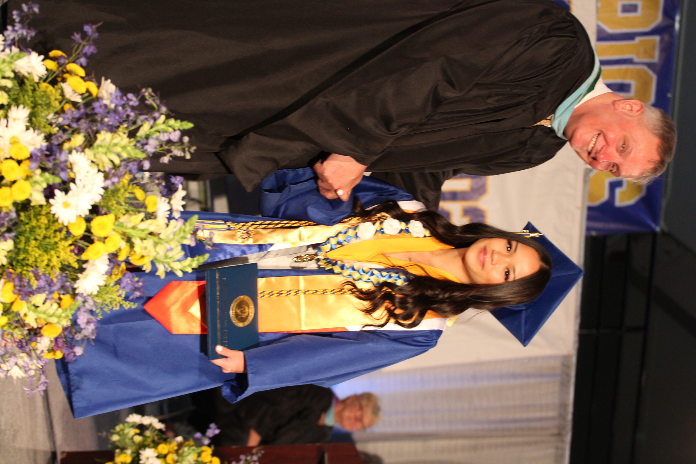 A man and a woman in academic gowns and caps on a stage with flowers. The man shakes the woman's hand.