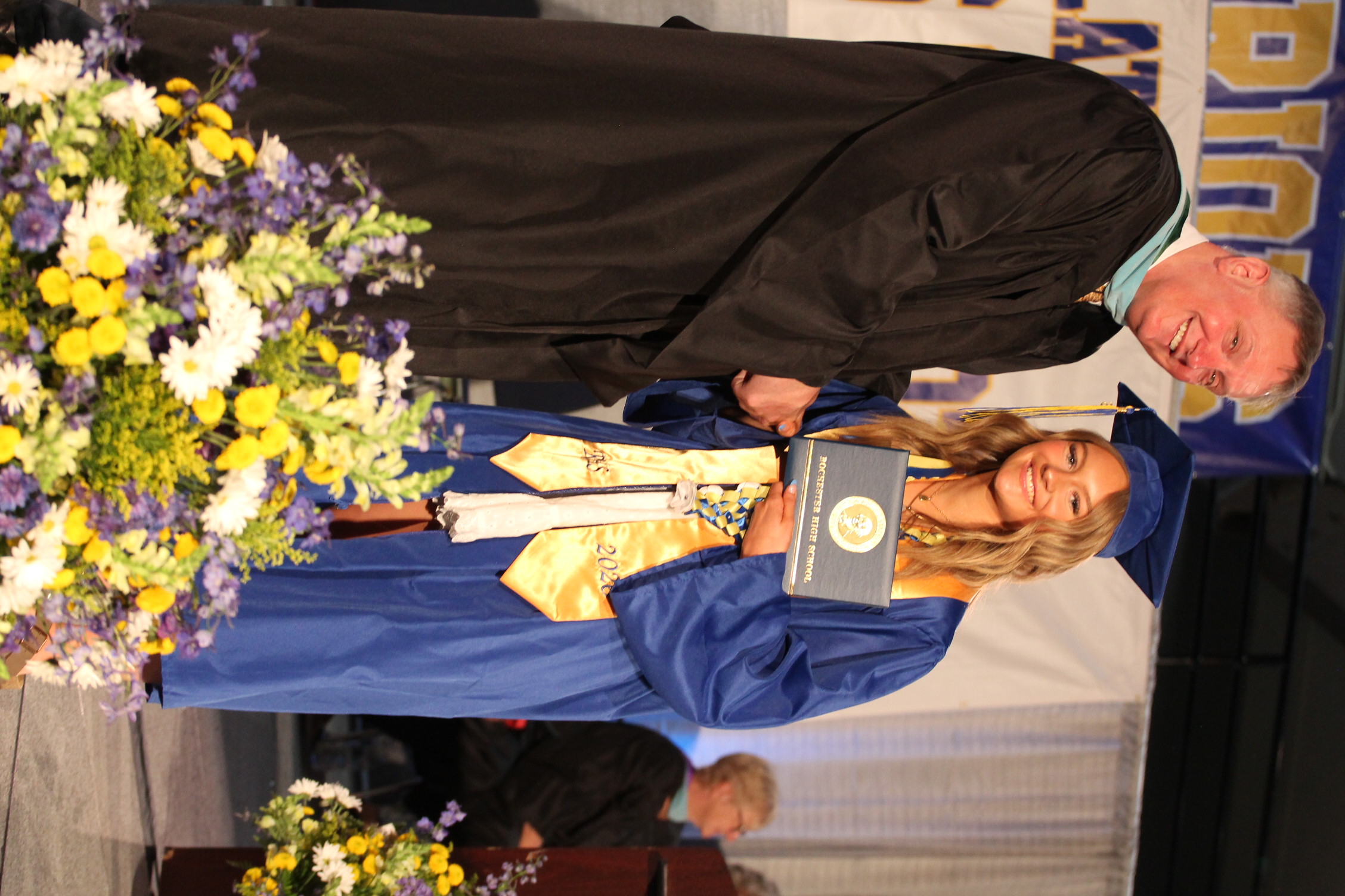 A male and female in graduation gowns stand on stage; the man hands the woman a diploma. Behind them, a banner reads "DIOS".
