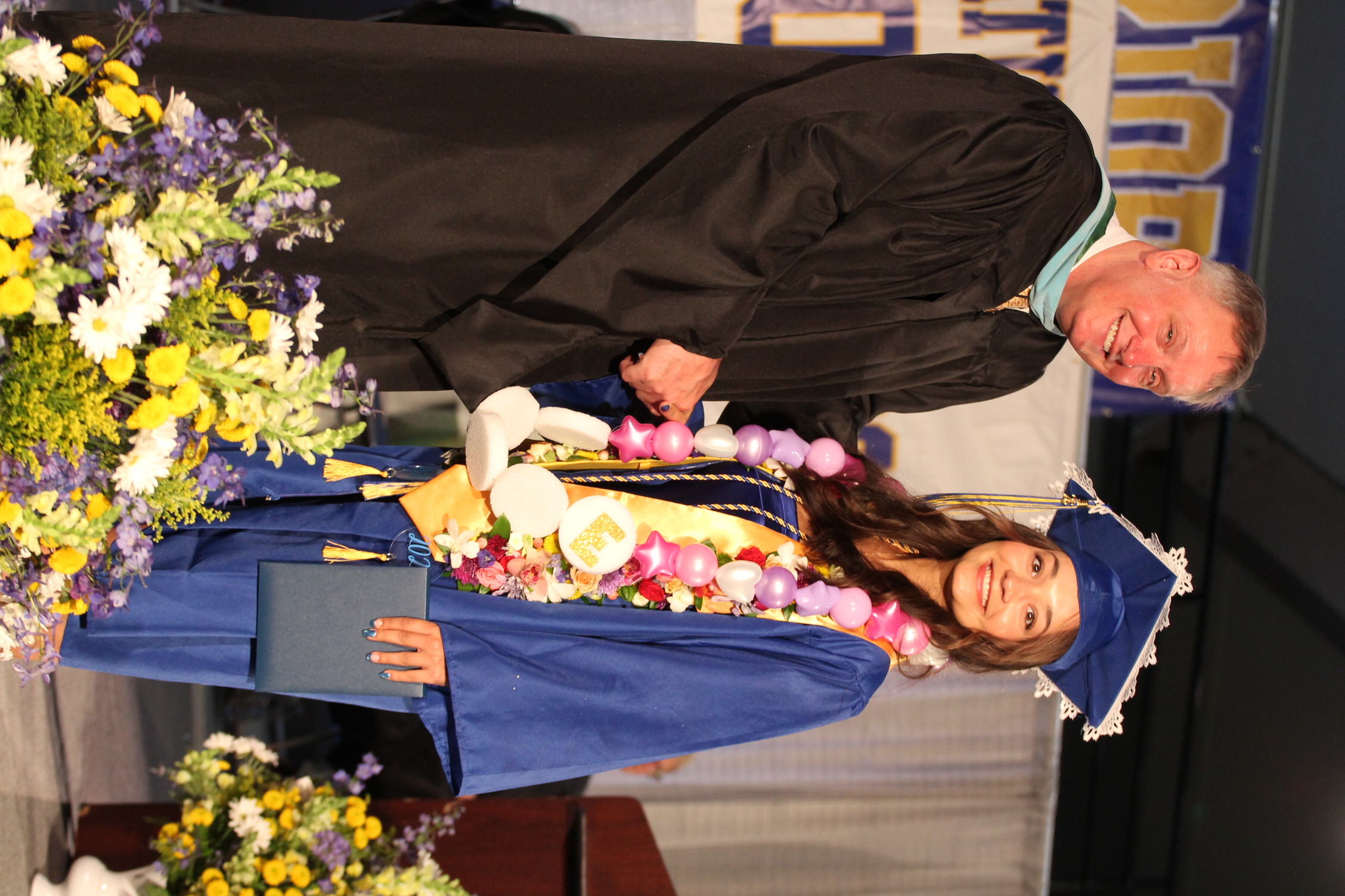 A man and a woman in graduation attire stand close together; the man smiles and holds a diploma.