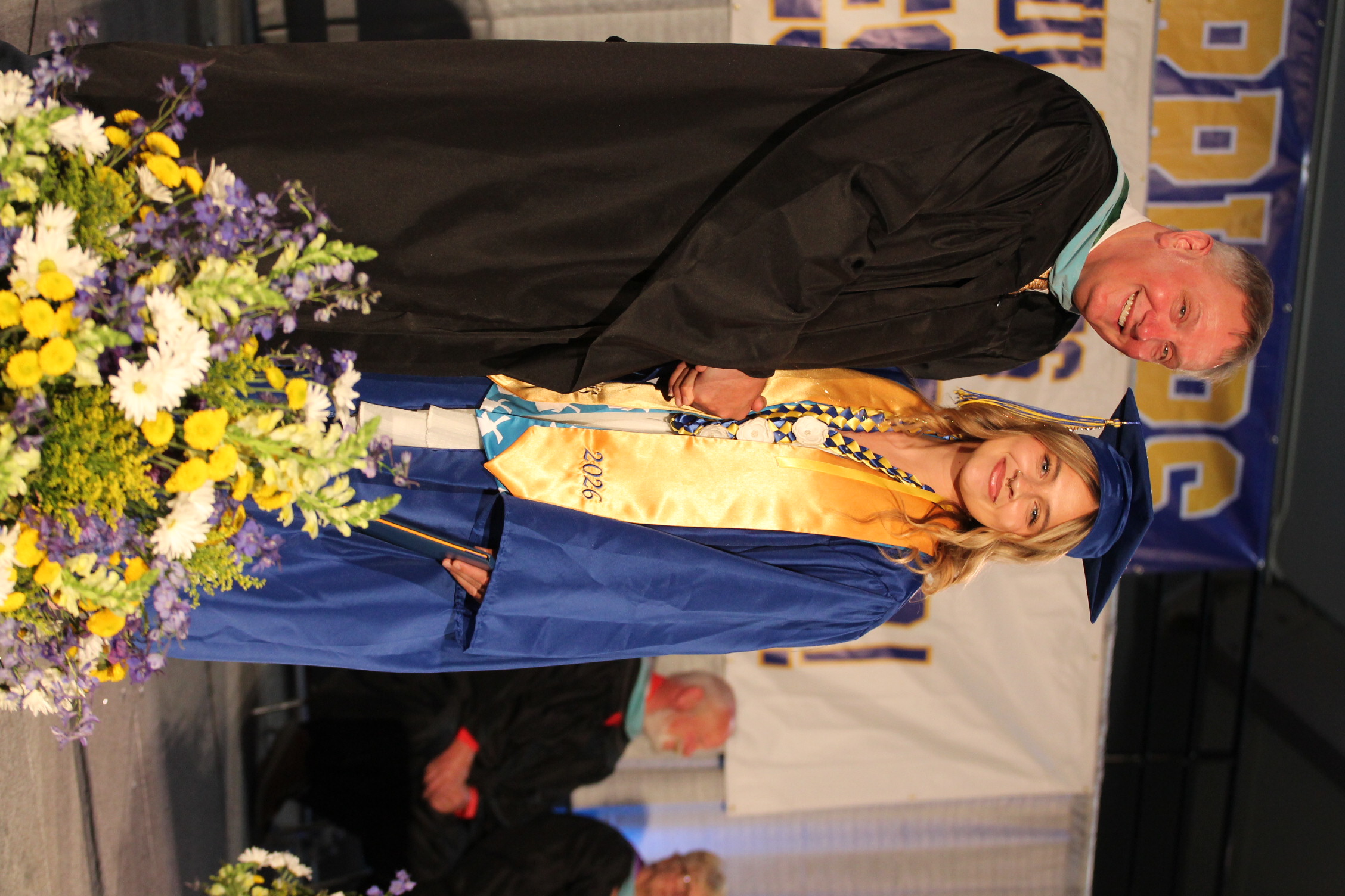 A smiling man and woman in graduation robes stand next to a flower arrangement on stage.