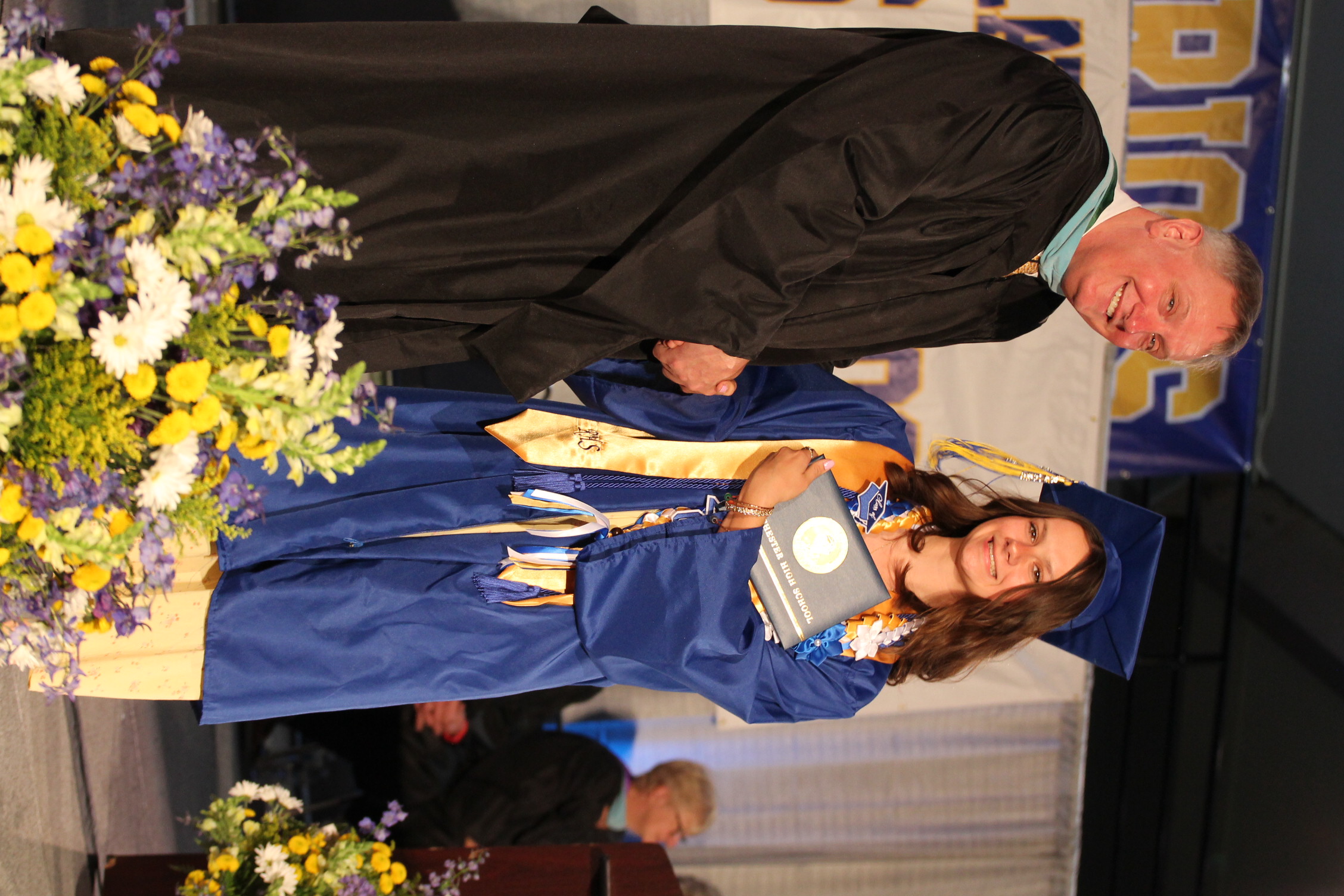 Man in black graduation robe shakes hand with woman in blue gown holding diploma. Flowers in foreground.