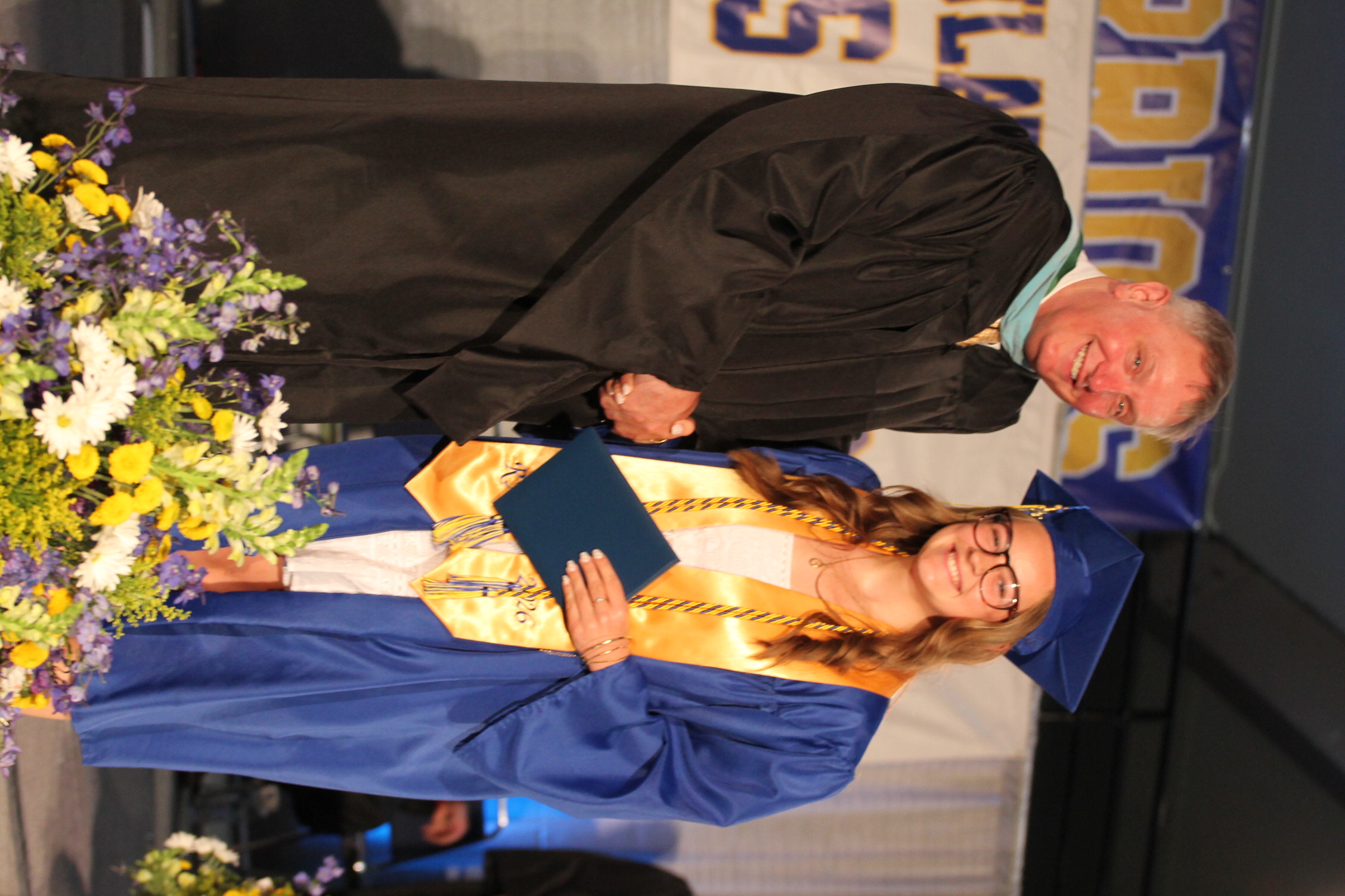 Man in black gown shakes a woman's hand. Woman in blue gown holds diploma, smiling. Floral arrangement on stage.