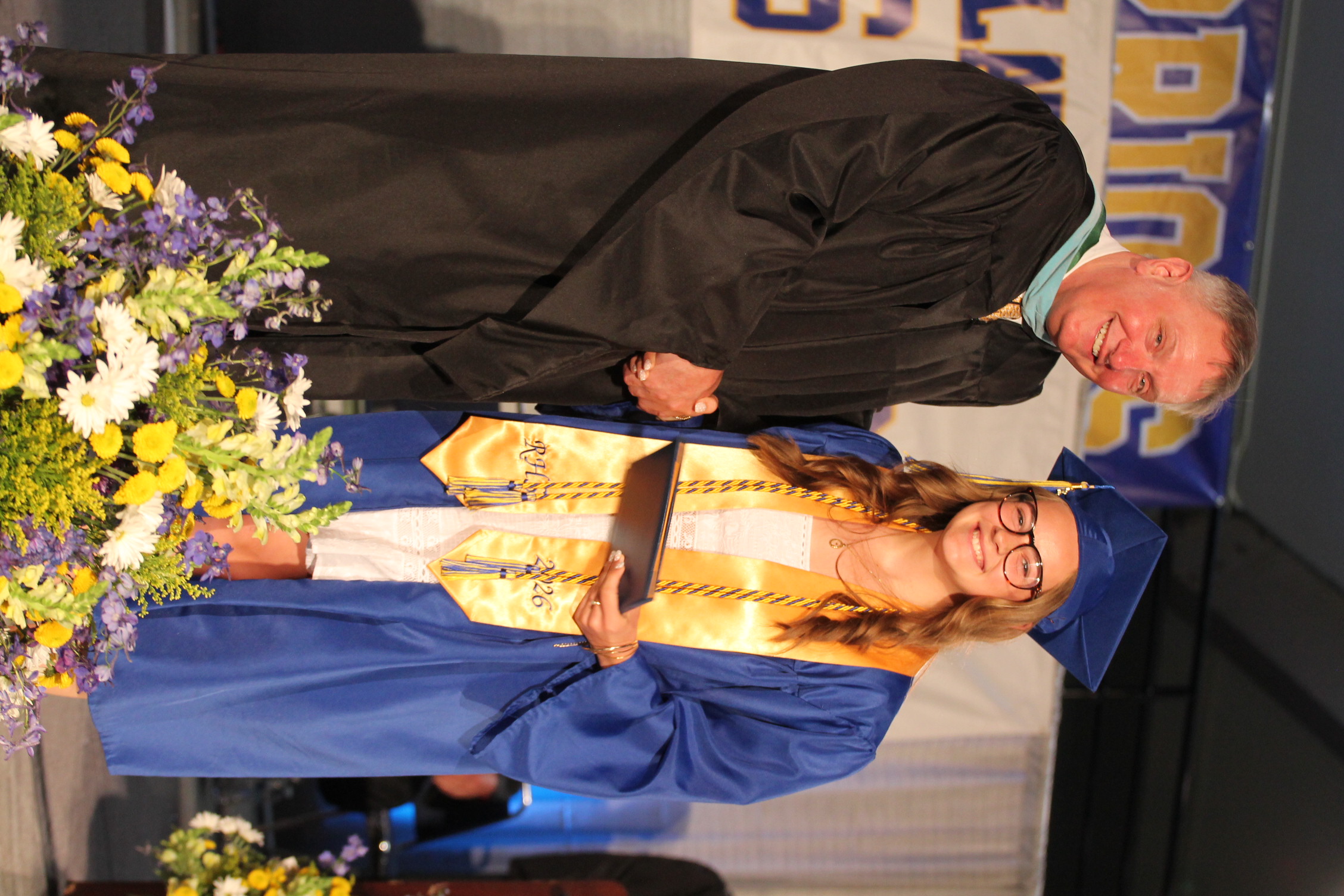 A man and woman on stage at a graduation ceremony. The woman in a blue gown holds a diploma.