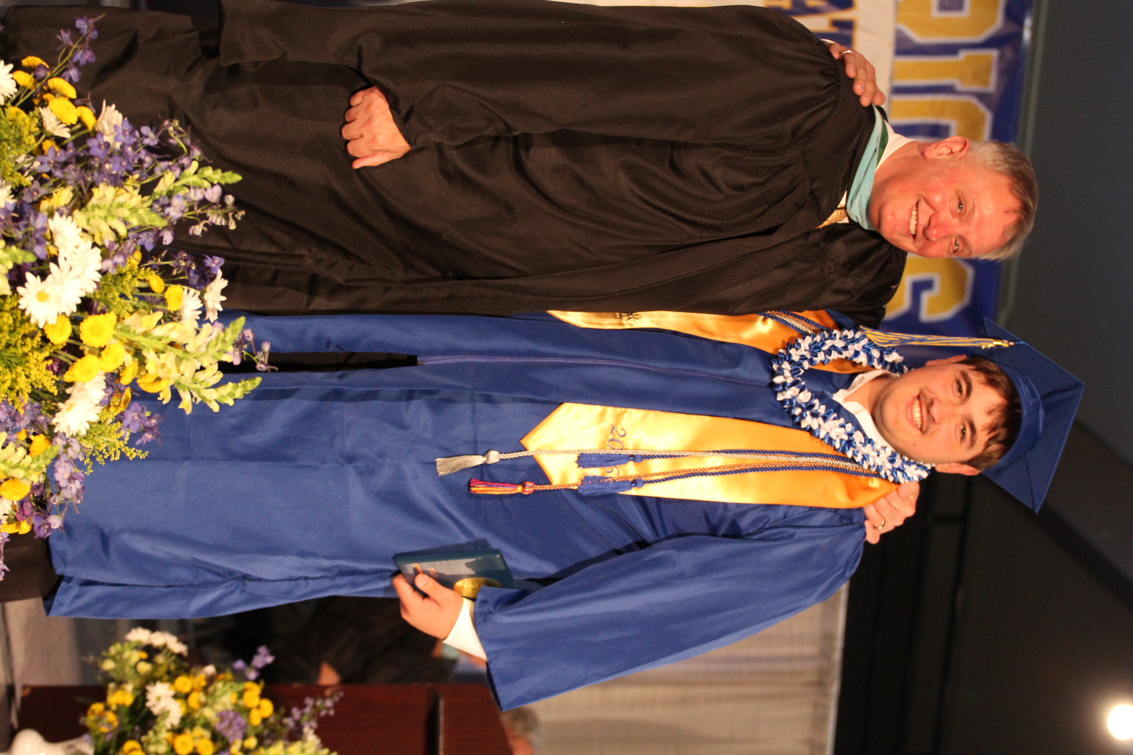 A man in a black gown and a man in a blue gown with a lei stand together with a flower arrangement.