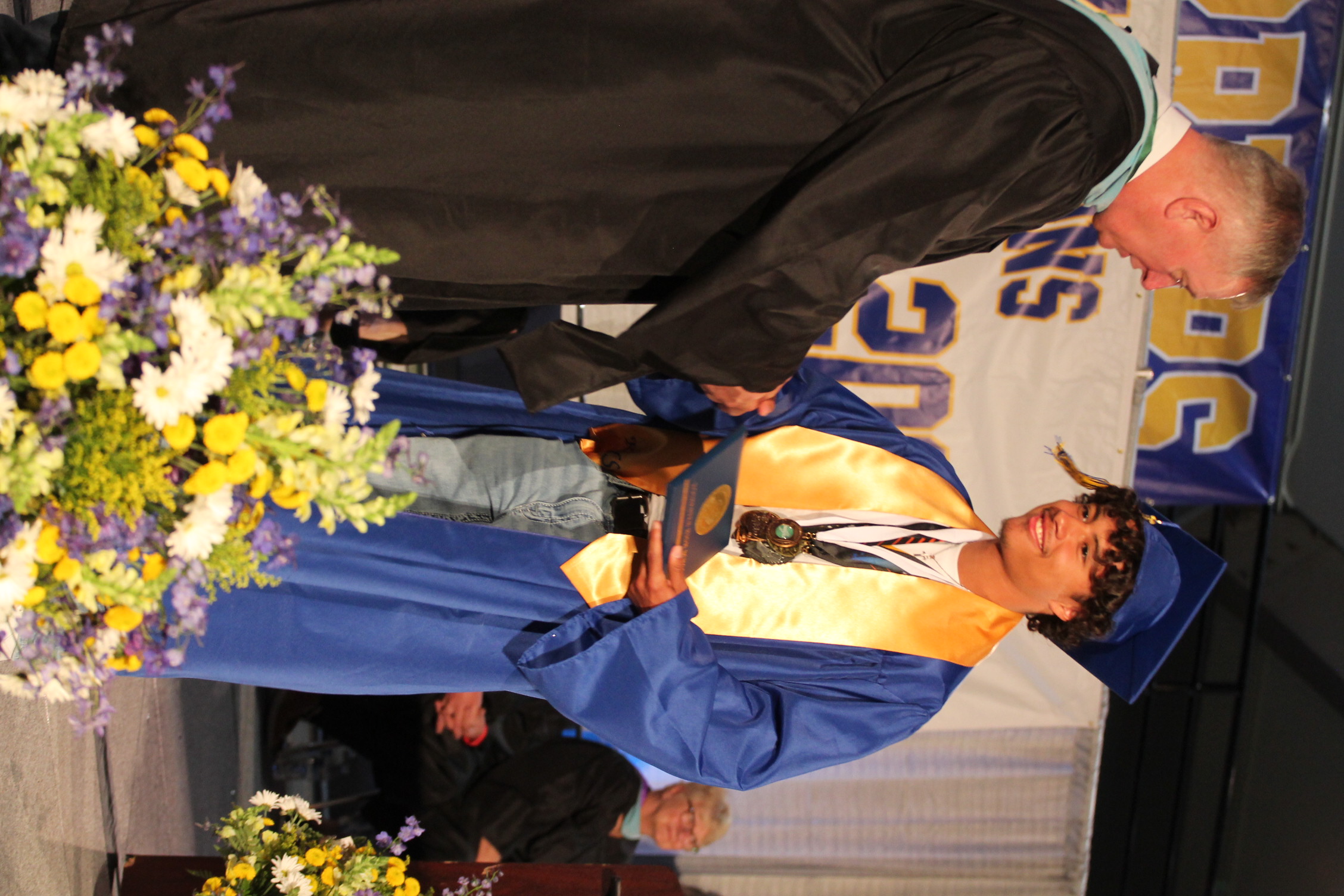 A man in a black gown hands a diploma to a graduate wearing a blue gown, yellow stole, and cap.